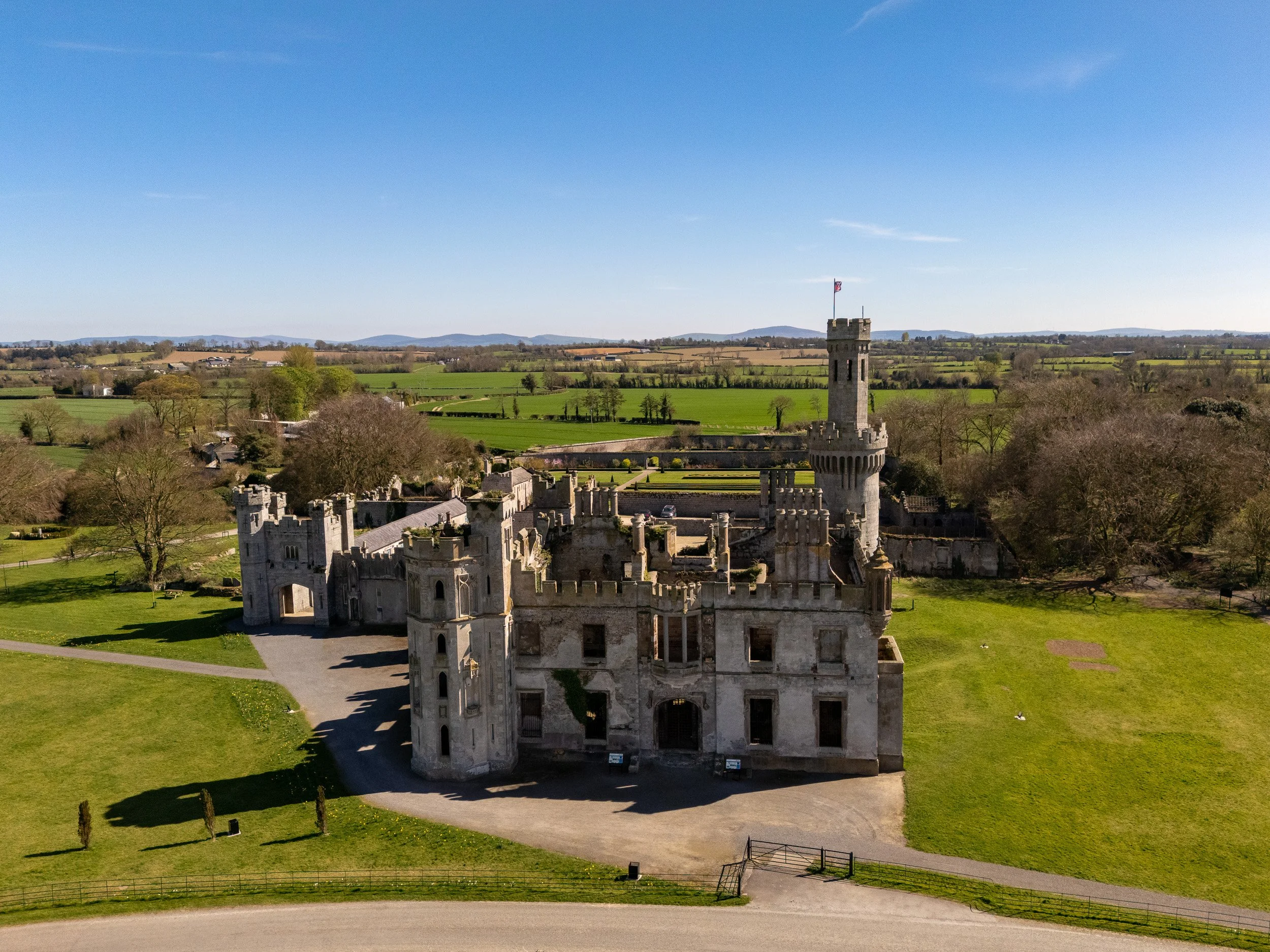 Aerial view of a historic castle with stone walls and towers, surrounded by green fields and trees under a clear blue sky.County Carlow.