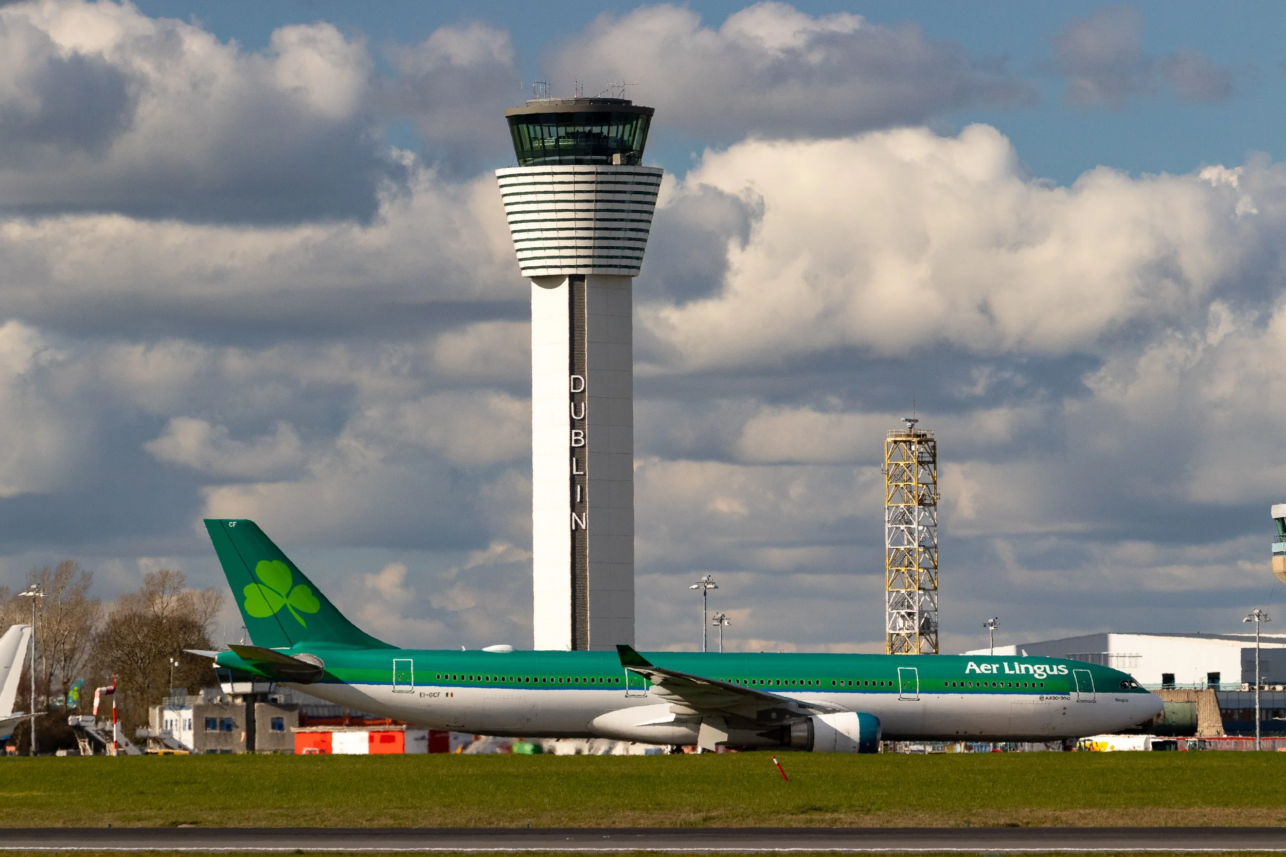An airplane painted in green and white with the words 'Aer Lingus' on the side, parked on the tarmac at  Dublin airport. In the background, there is a control tower and other airport structures under a partly cloudy sky.
