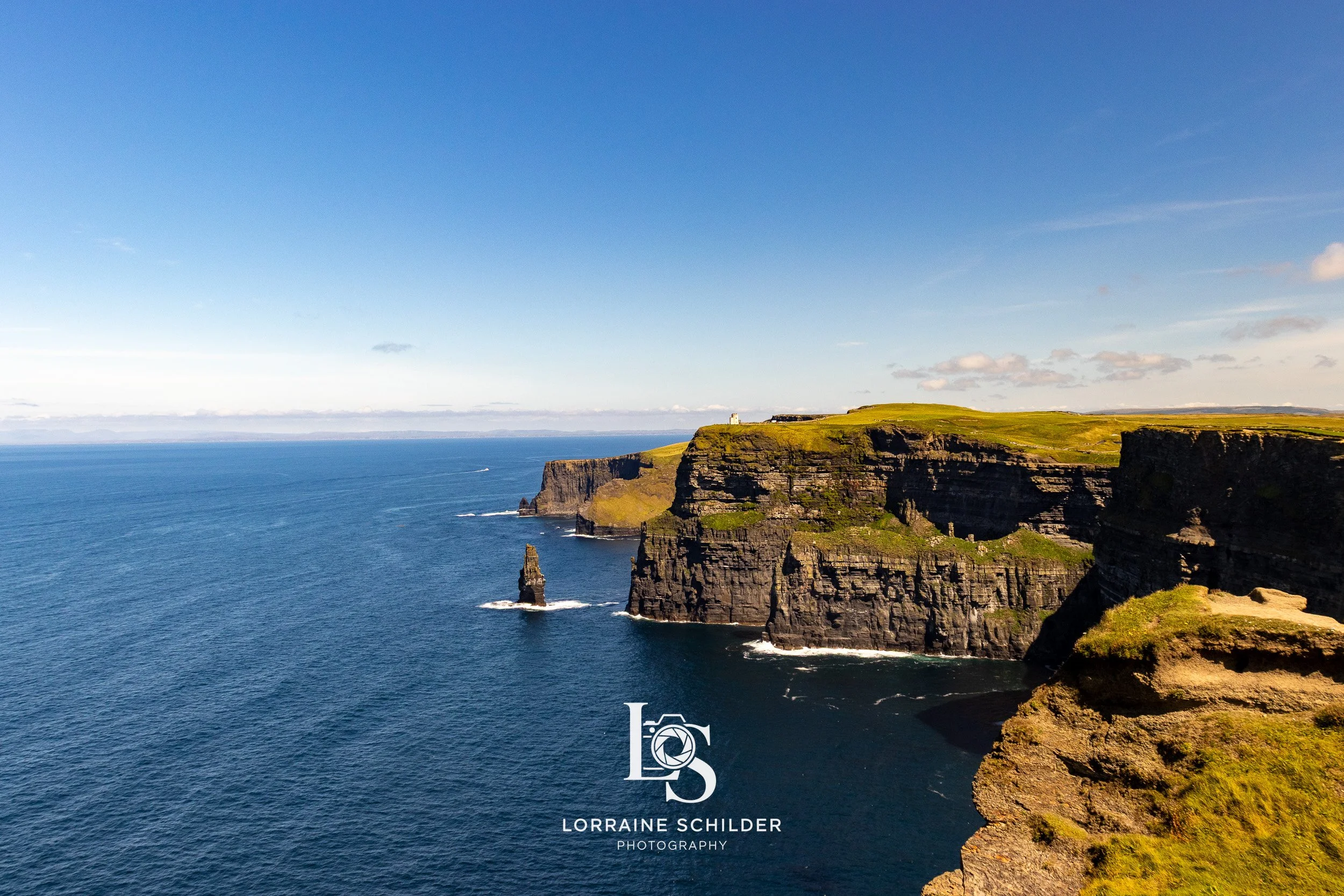 Cliffs overlooking the ocean with a clear blue sky, green grass on top, and a small white structure near the edge. Cliffs of Moher, Clare.