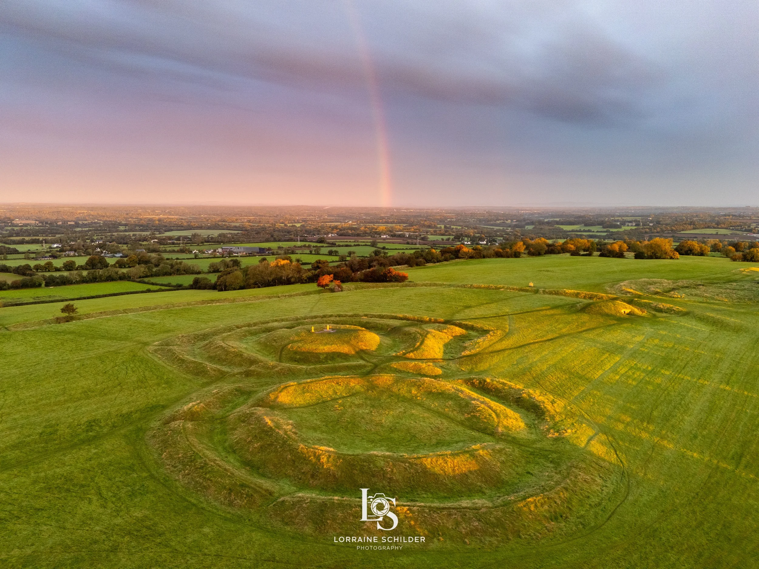 Aerial view of a green landscape with ancient stone circles, fields, trees, and a rainbow in the sky during sunrise.