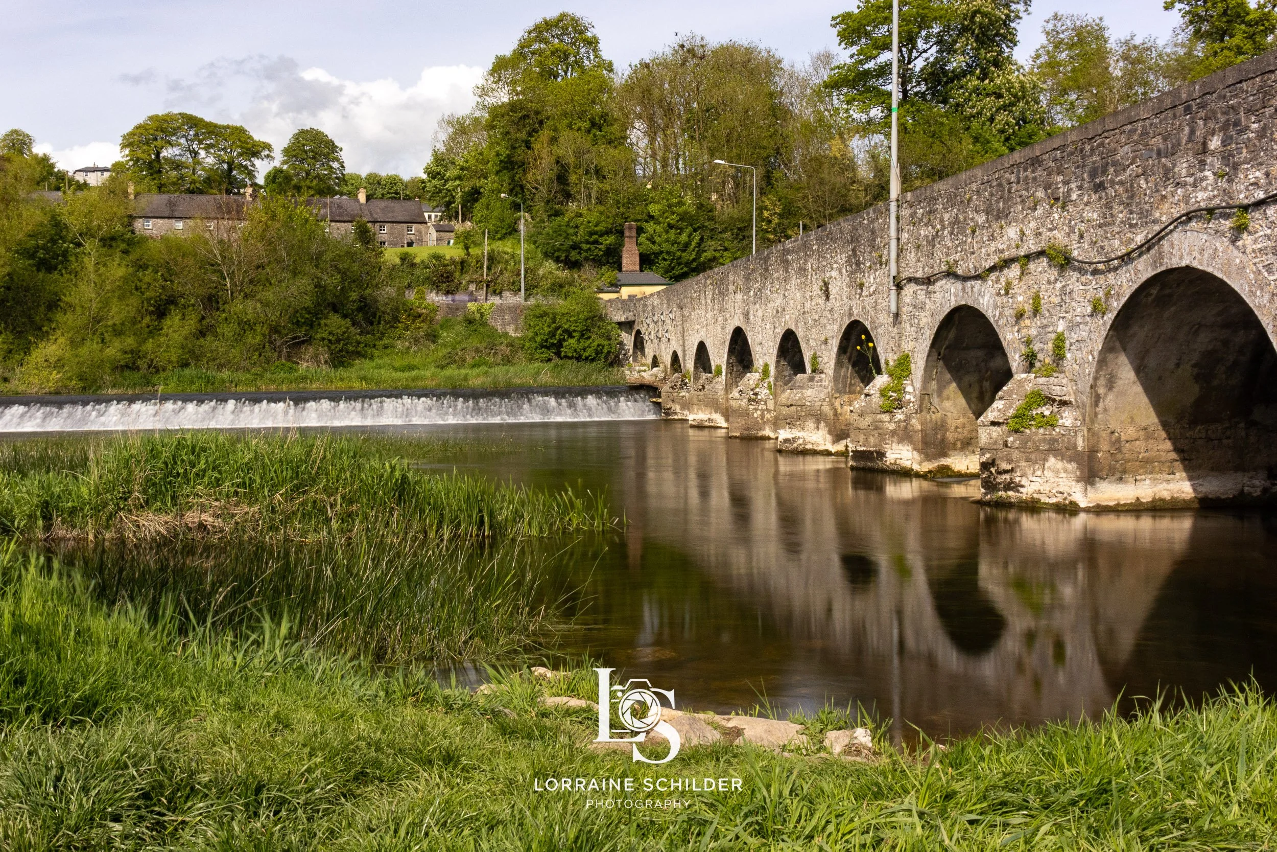 A stone arched bridge crossing a calm river with grass and trees on riverbanks, houses on a hill in background, and cloudy sky.  Slane, Meath