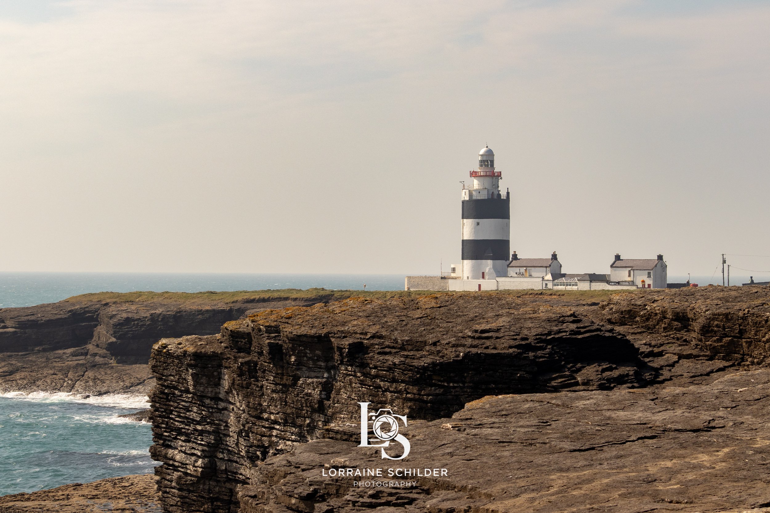 A black and white striped lighthouse on a rocky coastline with a cloudy sky and the ocean in the background. Wexford.