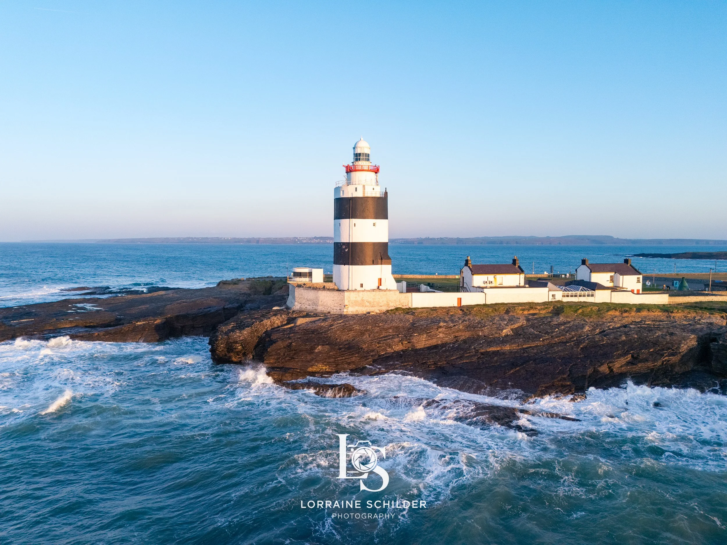 A lighthouse with black and white horizontal stripes standing on rocky coastline, with a few small white buildings nearby, under a clear blue sky and calm ocean waves. Wexford.