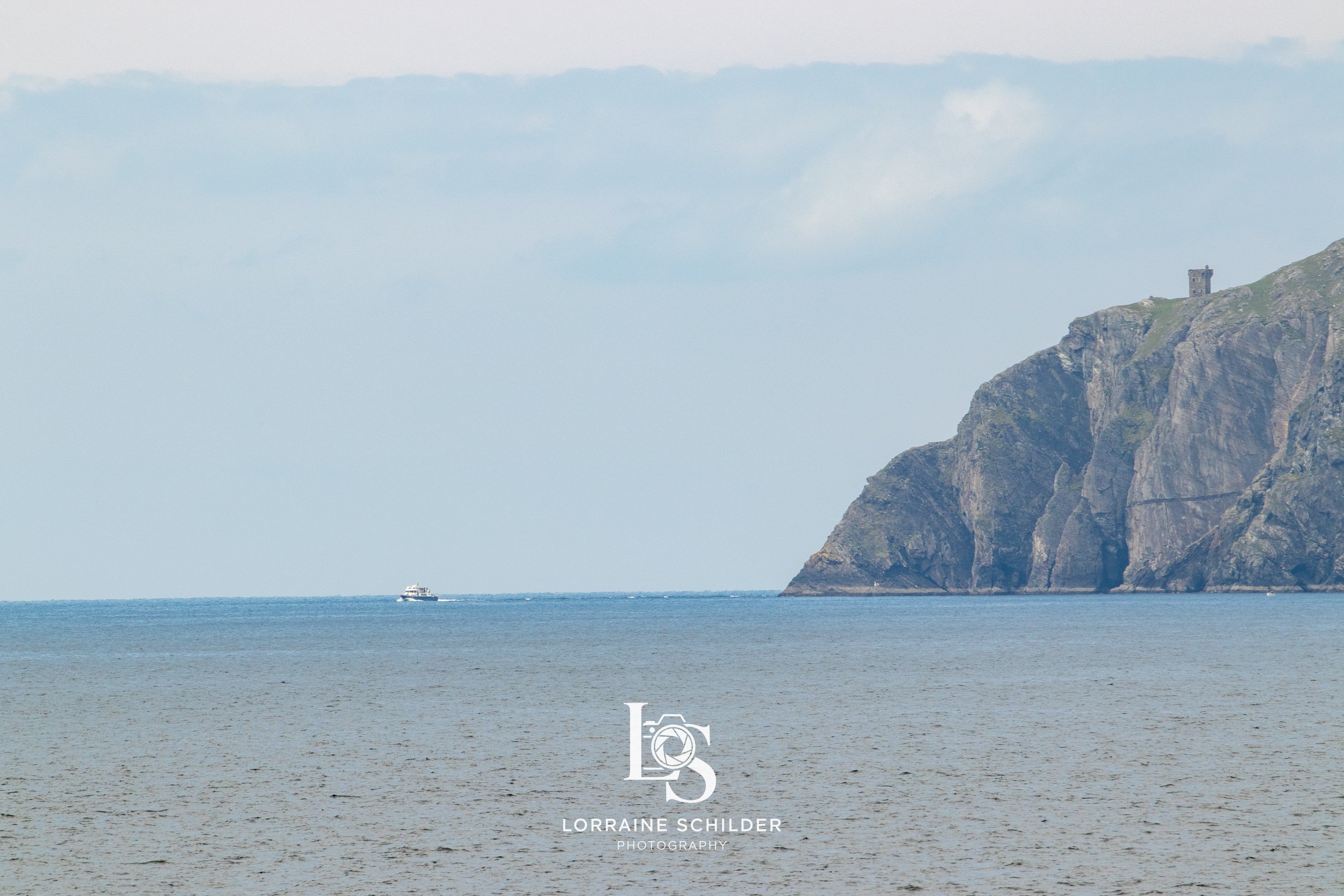 A seascape with a calm ocean, a small boat in the distance, and a rocky cliff with a castle on top under a cloudy sky.  Donegal.