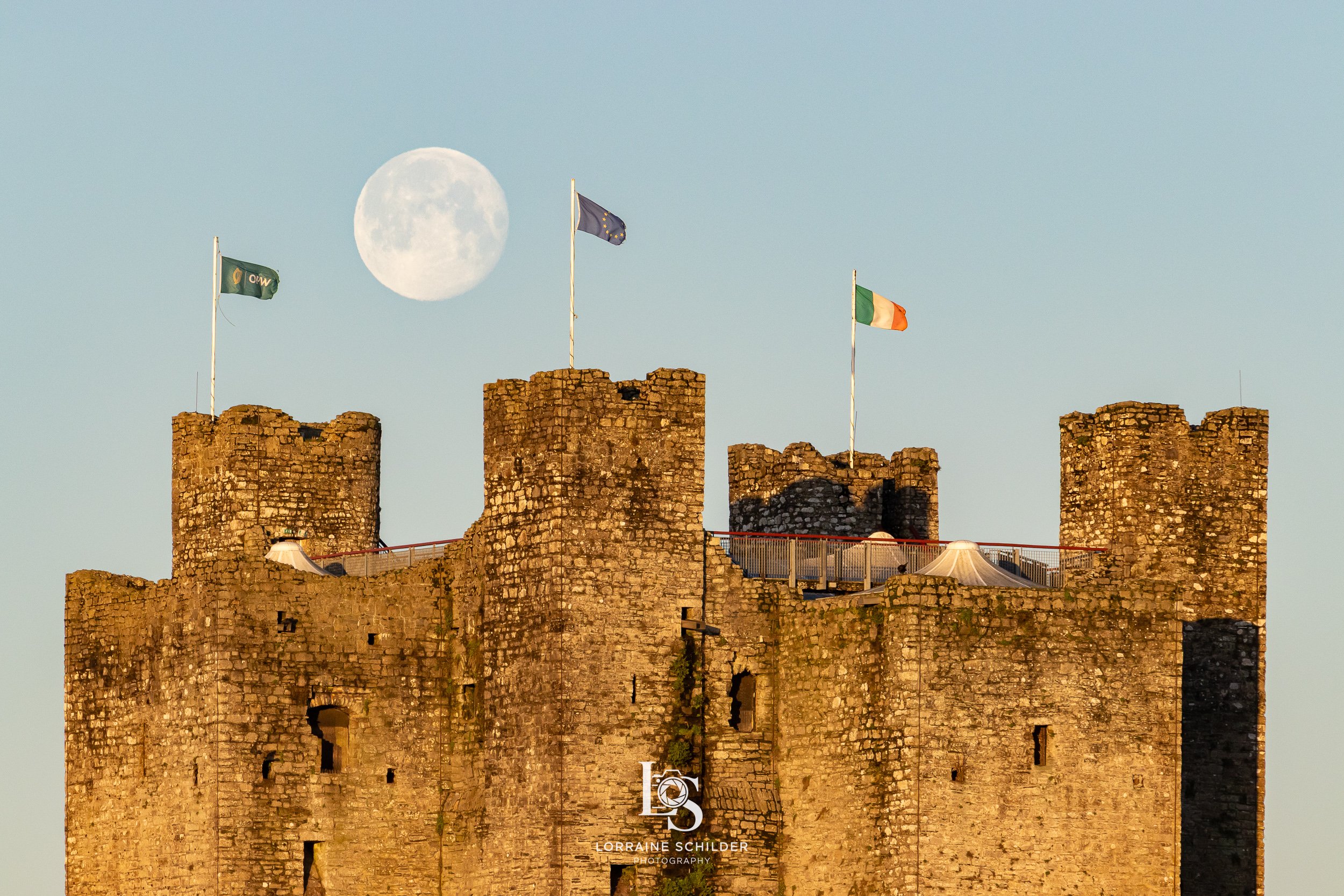 Trim Castle with flags on top, including the Irish flag, against a backdrop of a full moon and clear sky in the morning.