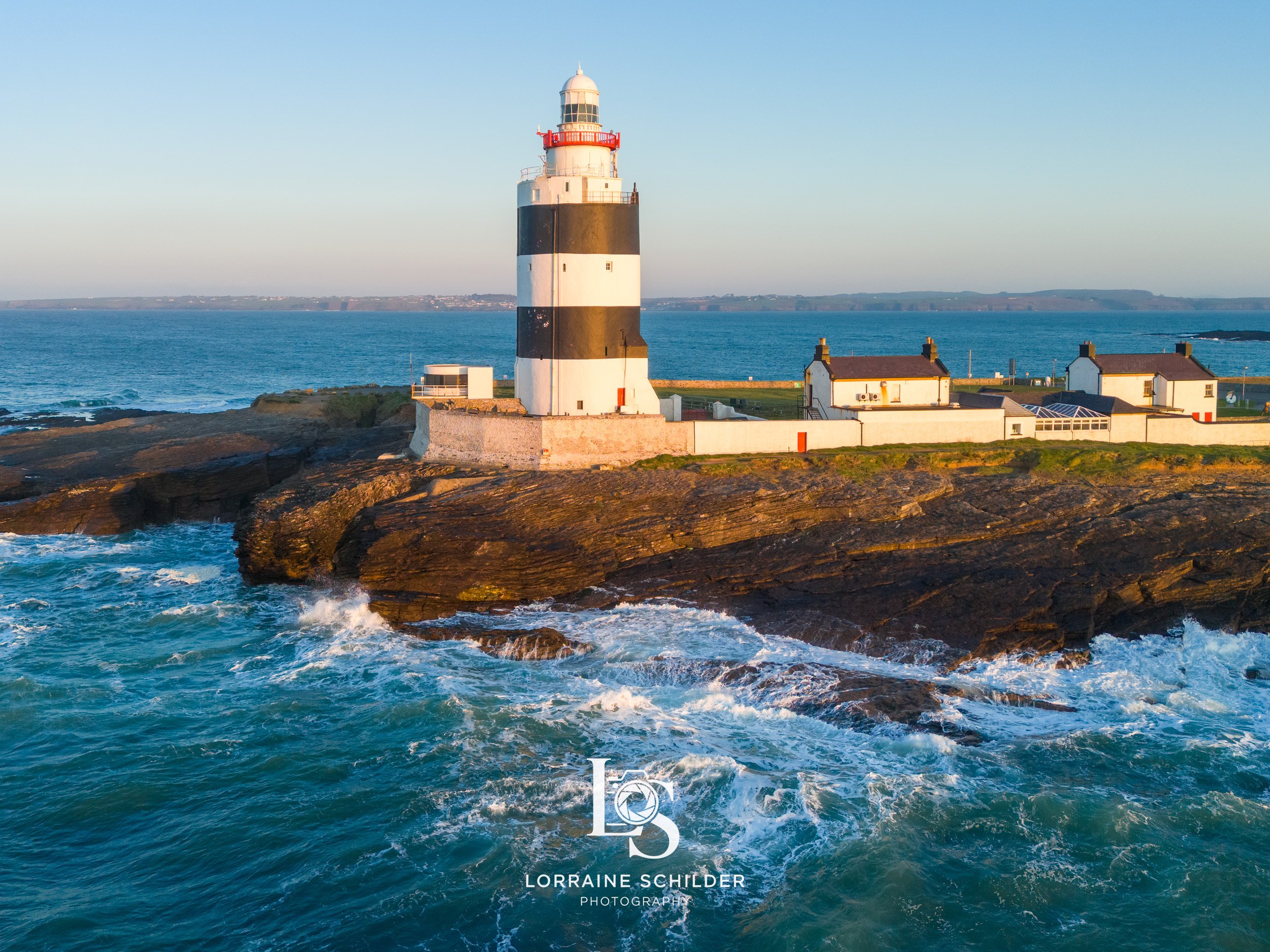 A lighthouse with black and white horizontal stripes on a rocky coastline with waves crashing, short grass, several small white houses with red chimneys, and the ocean in the background. Wexford.