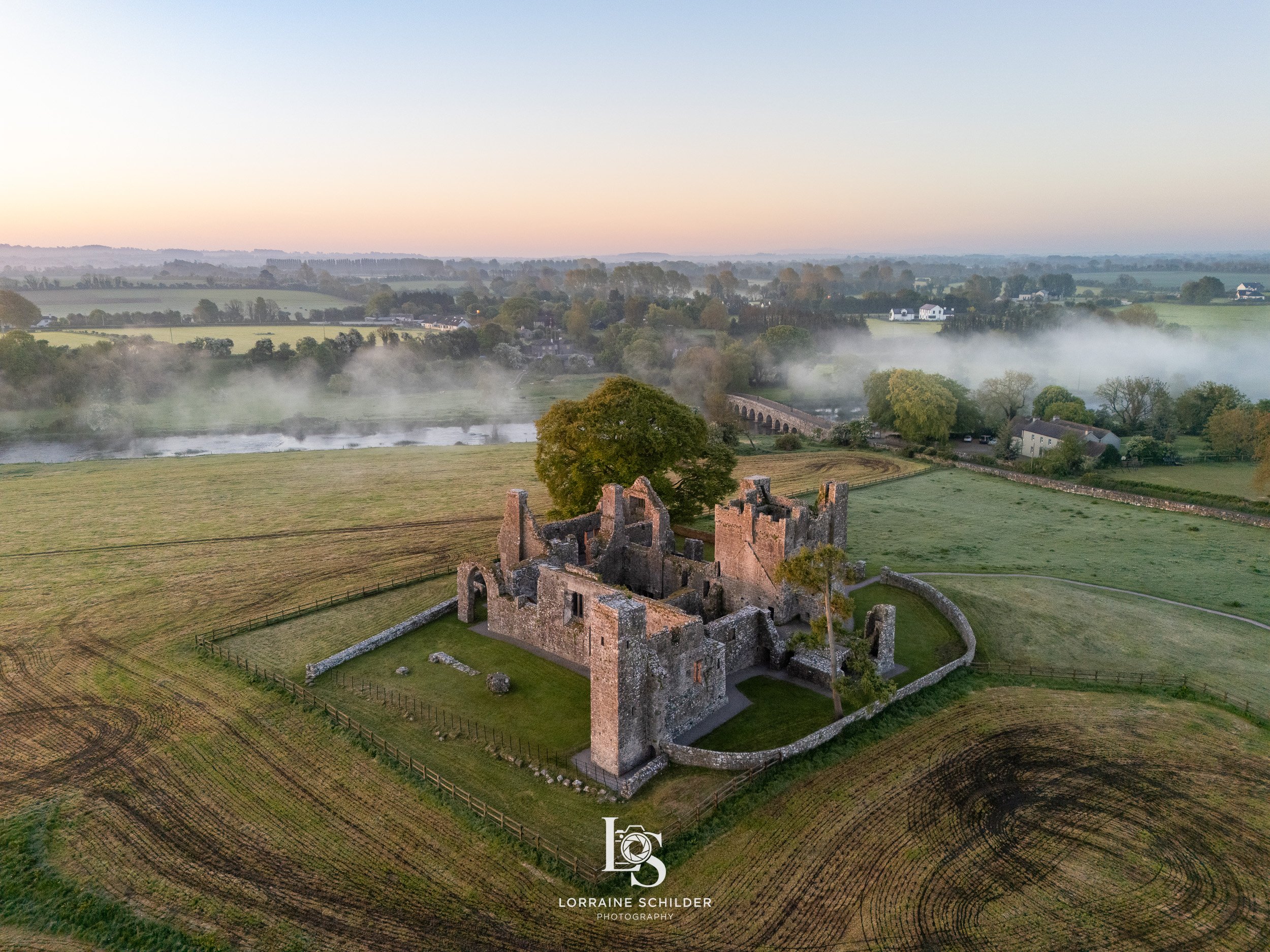 Aerial view of an ancient stone castle with surrounding green fields, trees, and a river in the background at sunrise or with fog over the landscape.  Bective Abbey, Meath.