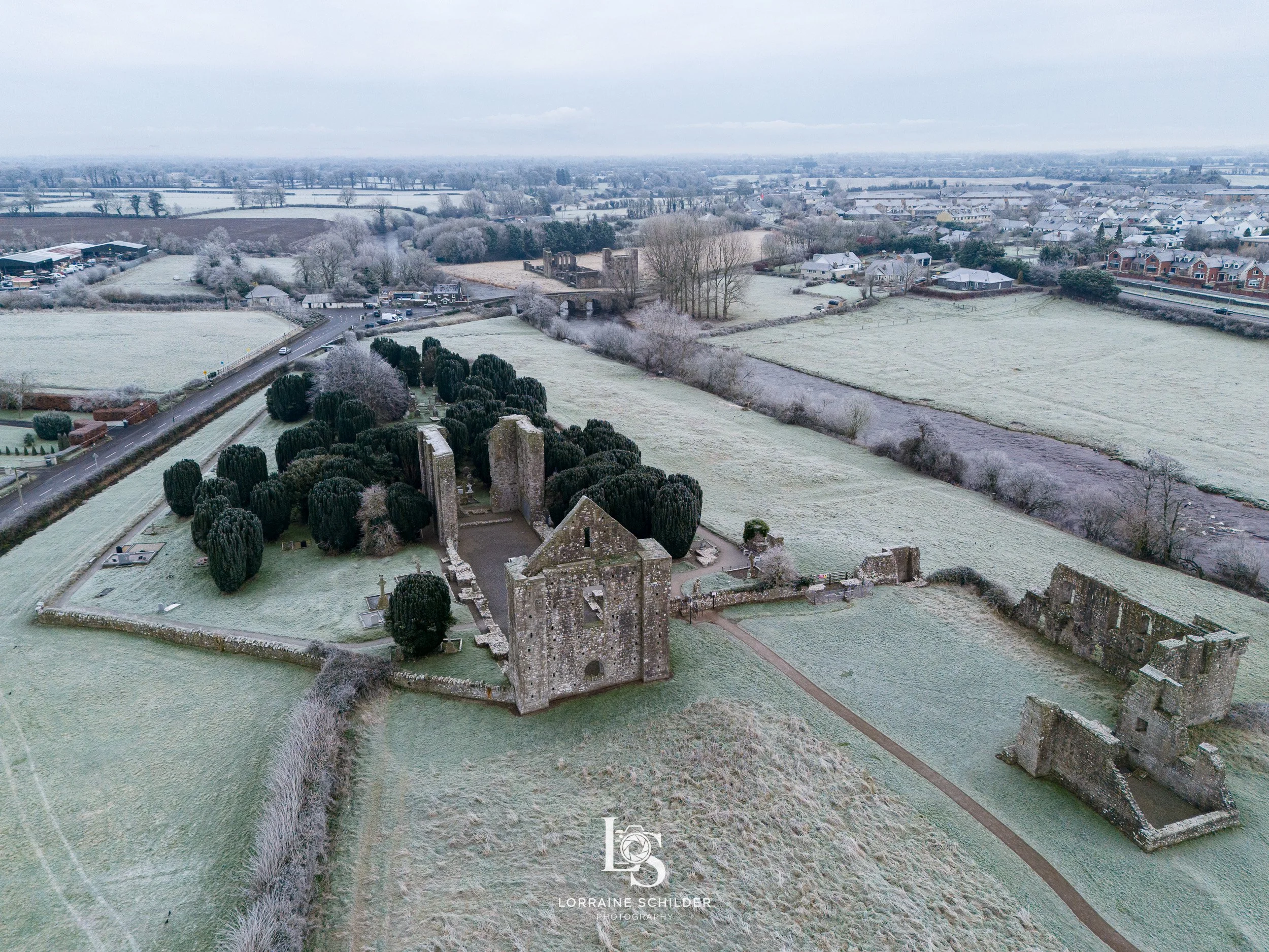 Aerial view Newtown Trim  in a green, frosty landscape with surrounding trees, old walls, and nearby farmland and residential area.