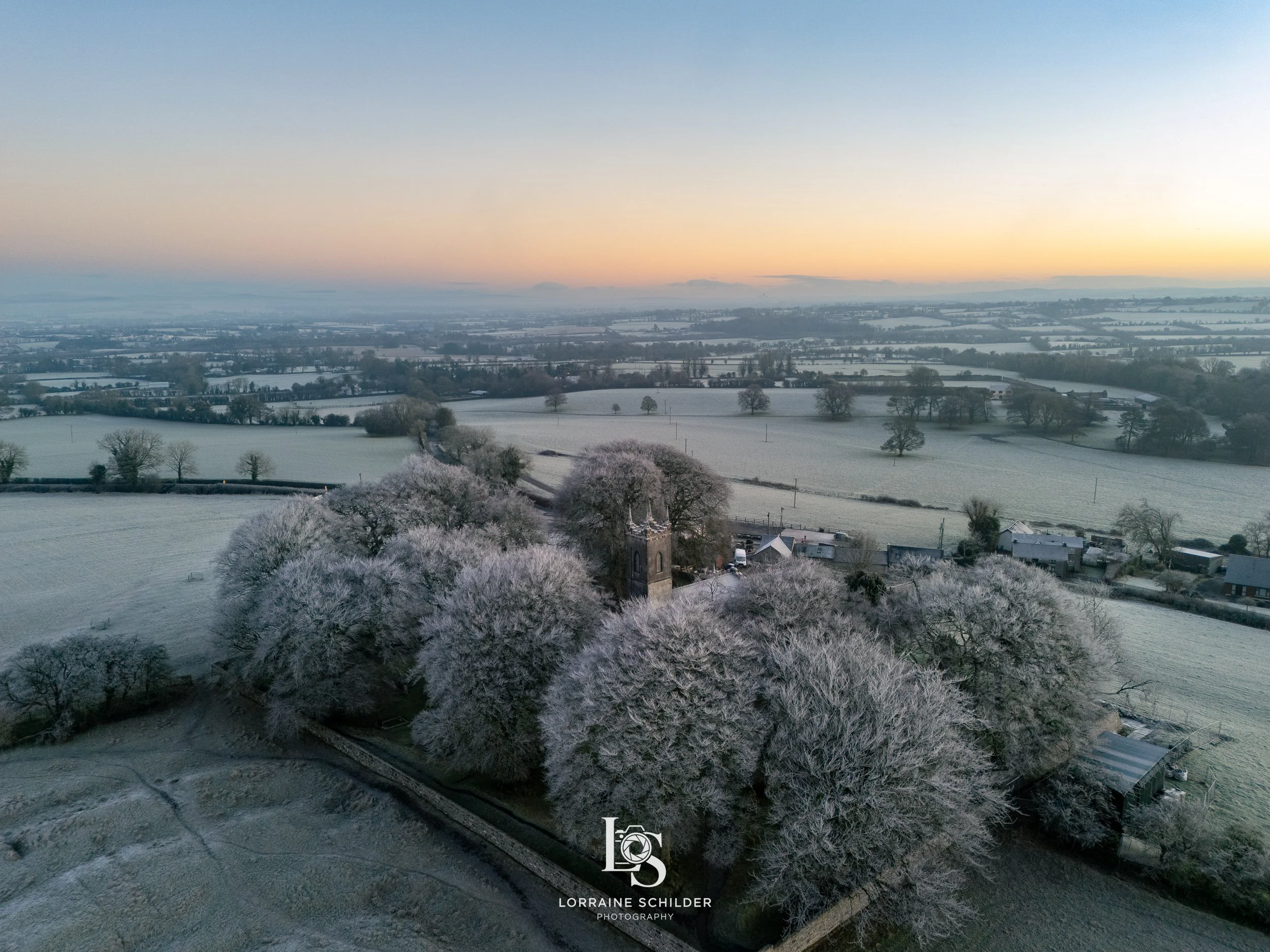 A winter landscape showing snow-covered fields and trees with a small village and church. The sky is clear with a subtle gradient from blue to orange near sunrise.
