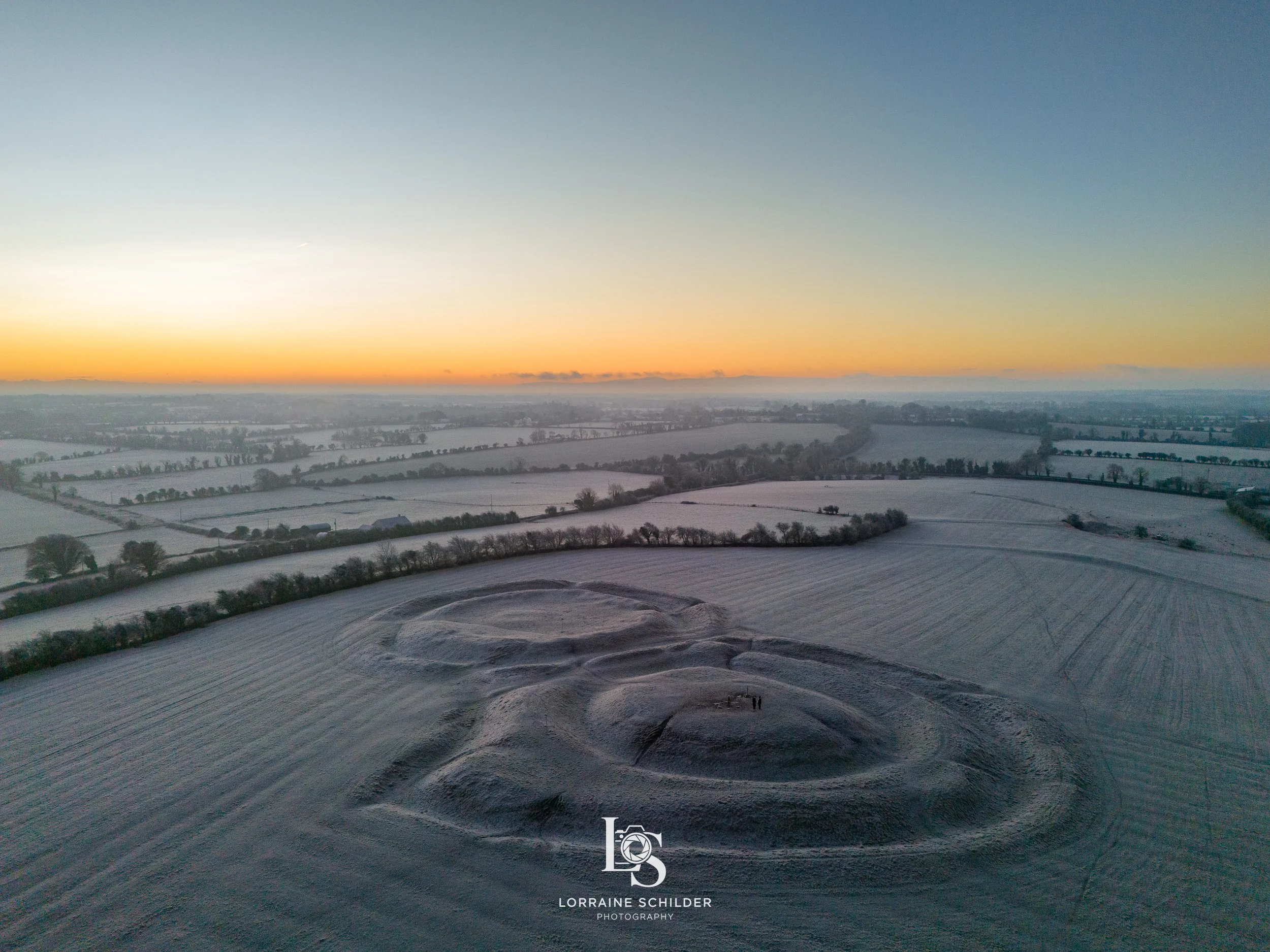 Snow-covered fields and a hill with a spiral pattern, at sunrise with a clear sky and distant horizon.