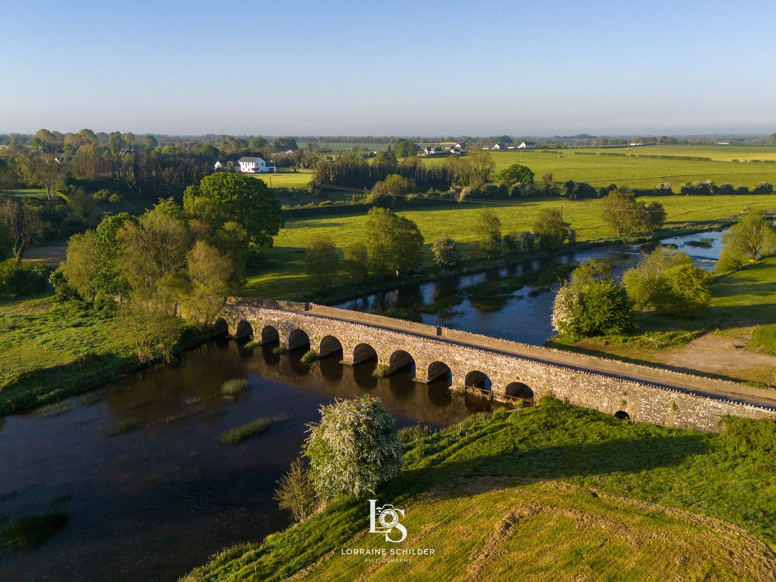 A scenic countryside view with a stone bridge over a winding river, surrounded by green fields and trees, under a clear blue sky.  Bective Bridge, Bective Abbey, Meath.