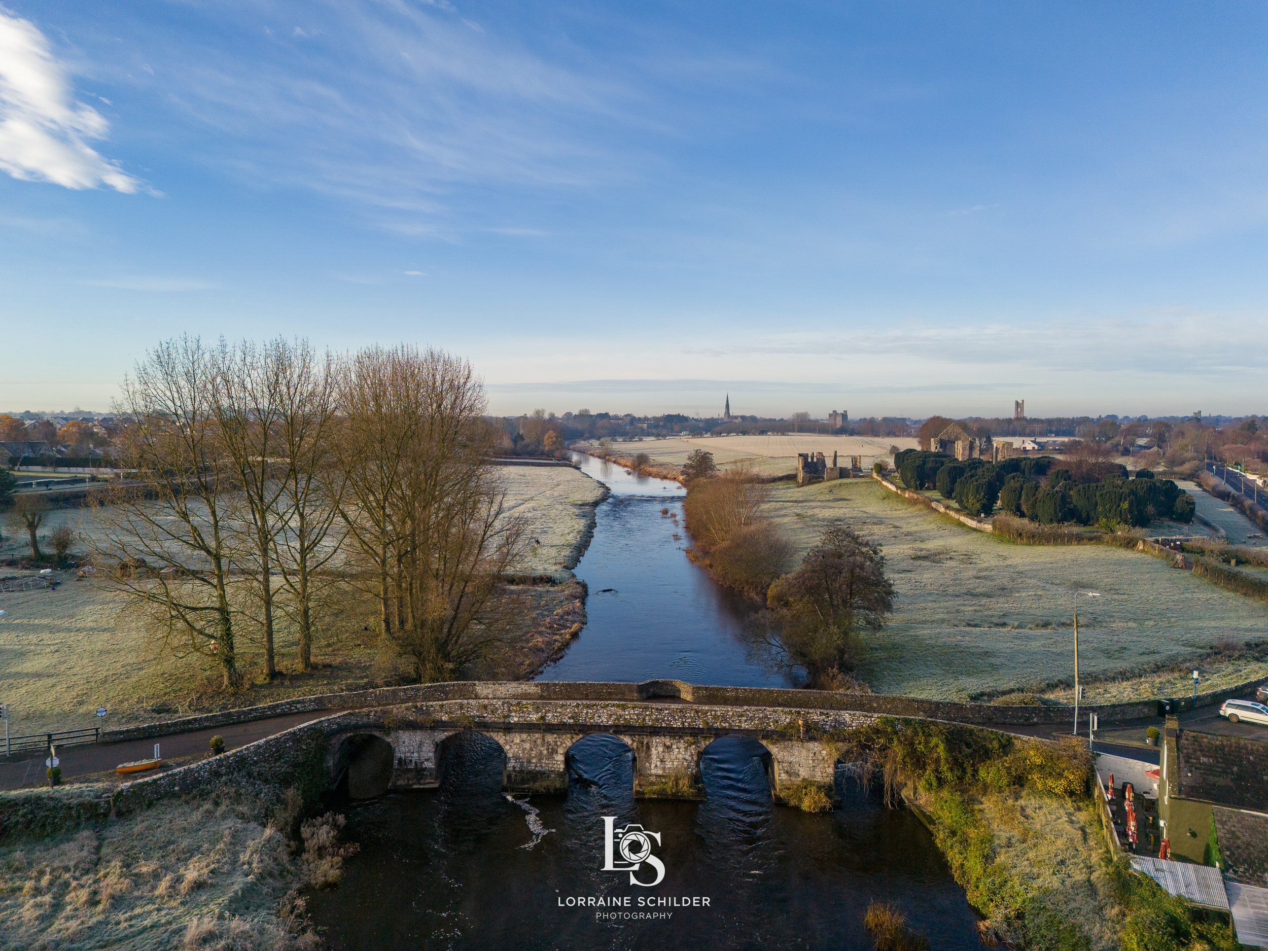 A scenic winter landscape featuring Newtown bridge over the Boyne River, leafless trees, grassy fields, and a distant church spire under a partly cloudy blue sky. Trim, meath.