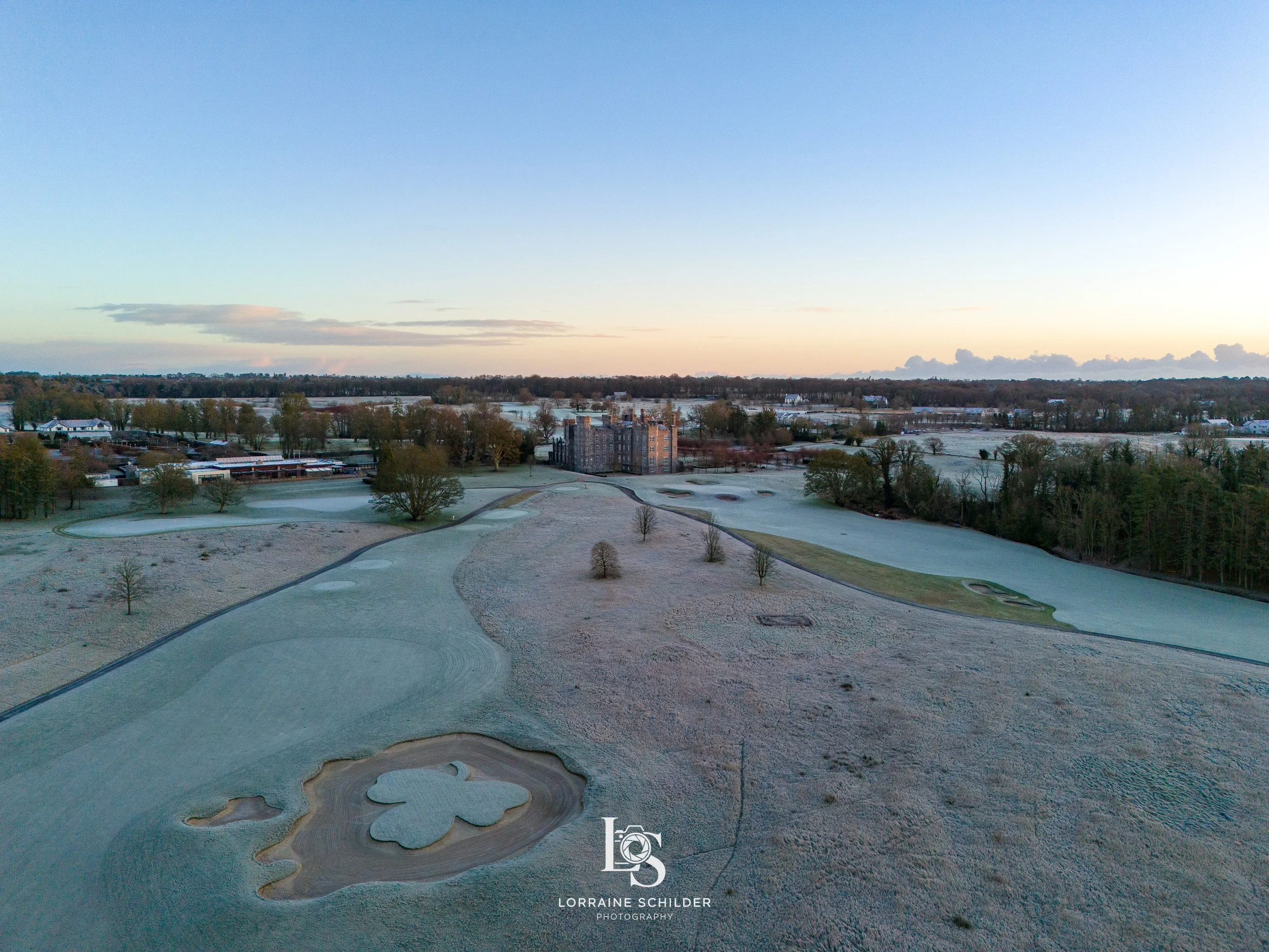 Aerial view of a golf course covered in frost, with several holes visible, trees, and a large building in the background under a clear sky at sunrise. Killeen Castle, Meath.