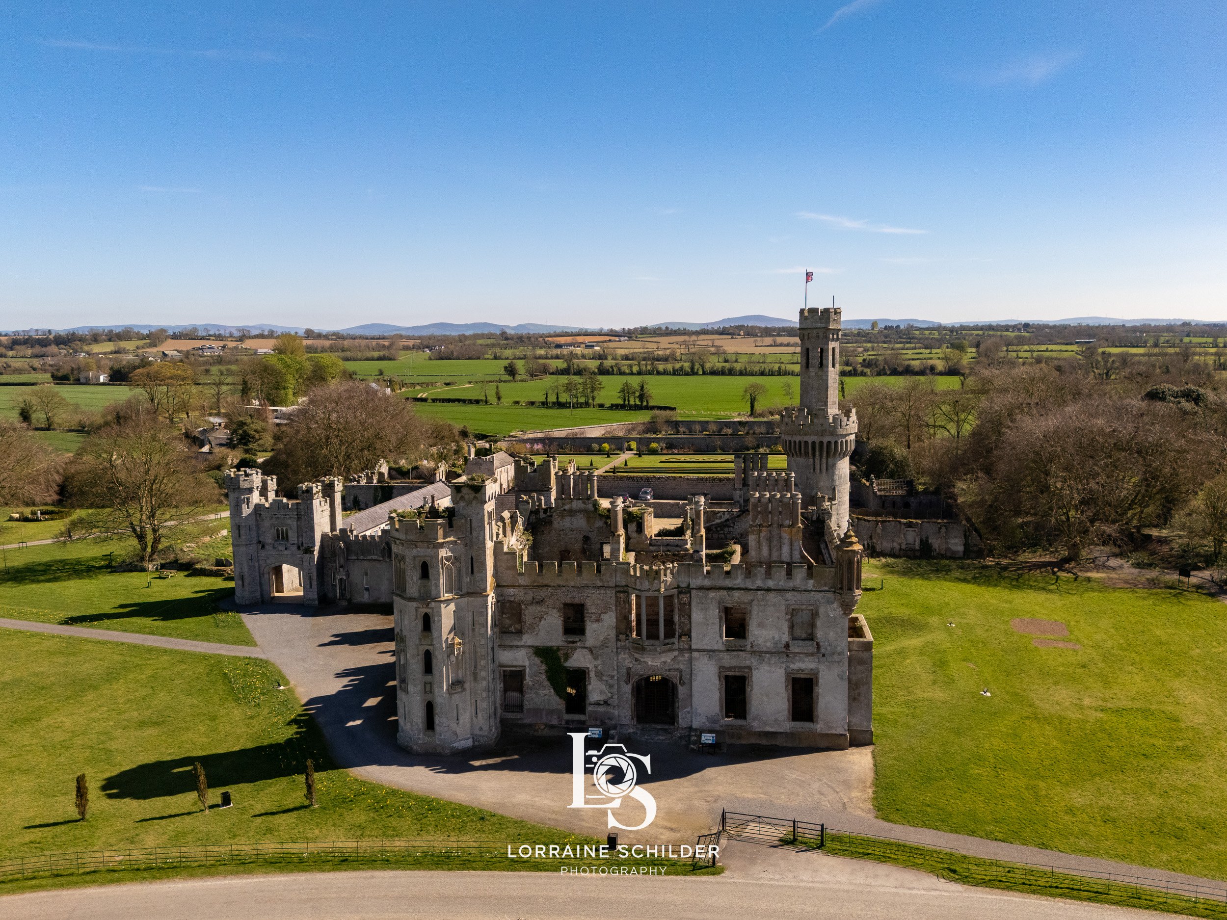 Aerial view of a historic castle with towers and a flag, surrounded by green fields and trees on a clear day. Carlow.