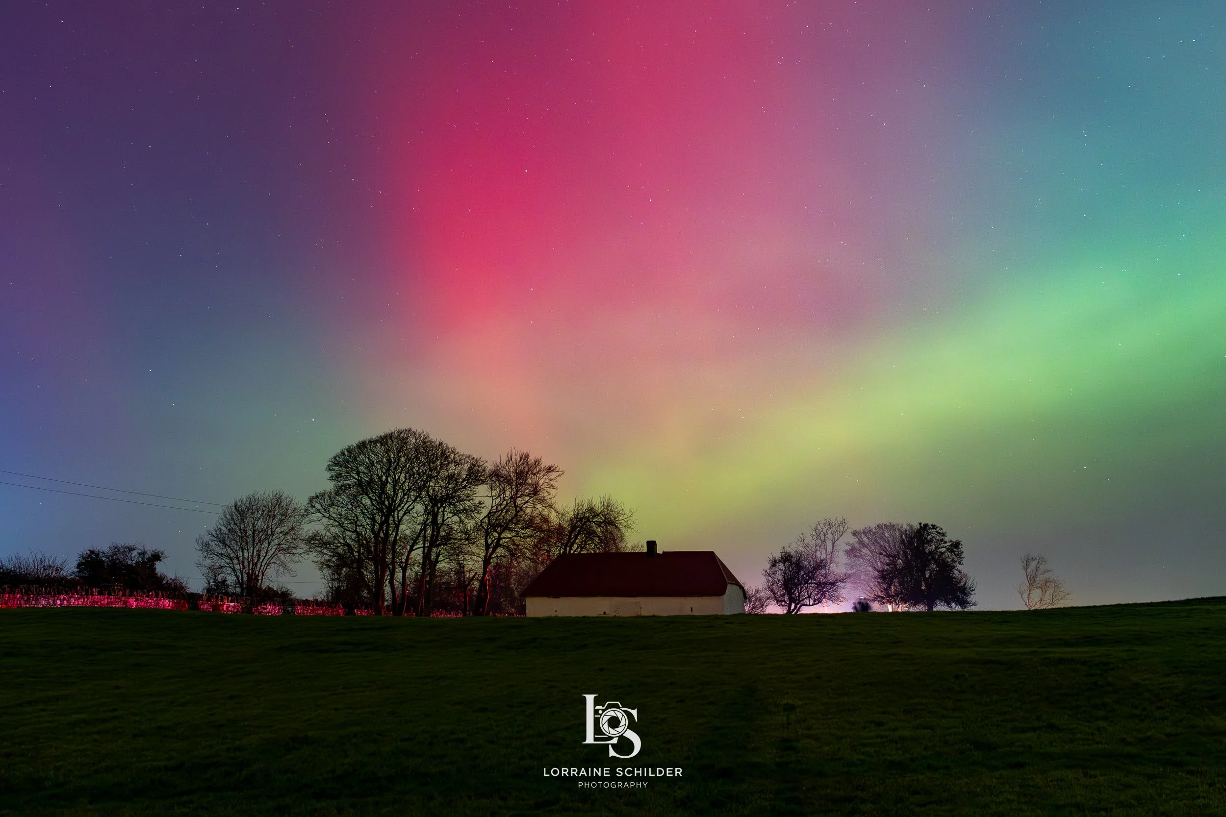 Nighttime scene with a colorful aurora borealis sky over a rural landscape. Silhouettes of leafless trees and a small house are visible against the vibrant sky.
