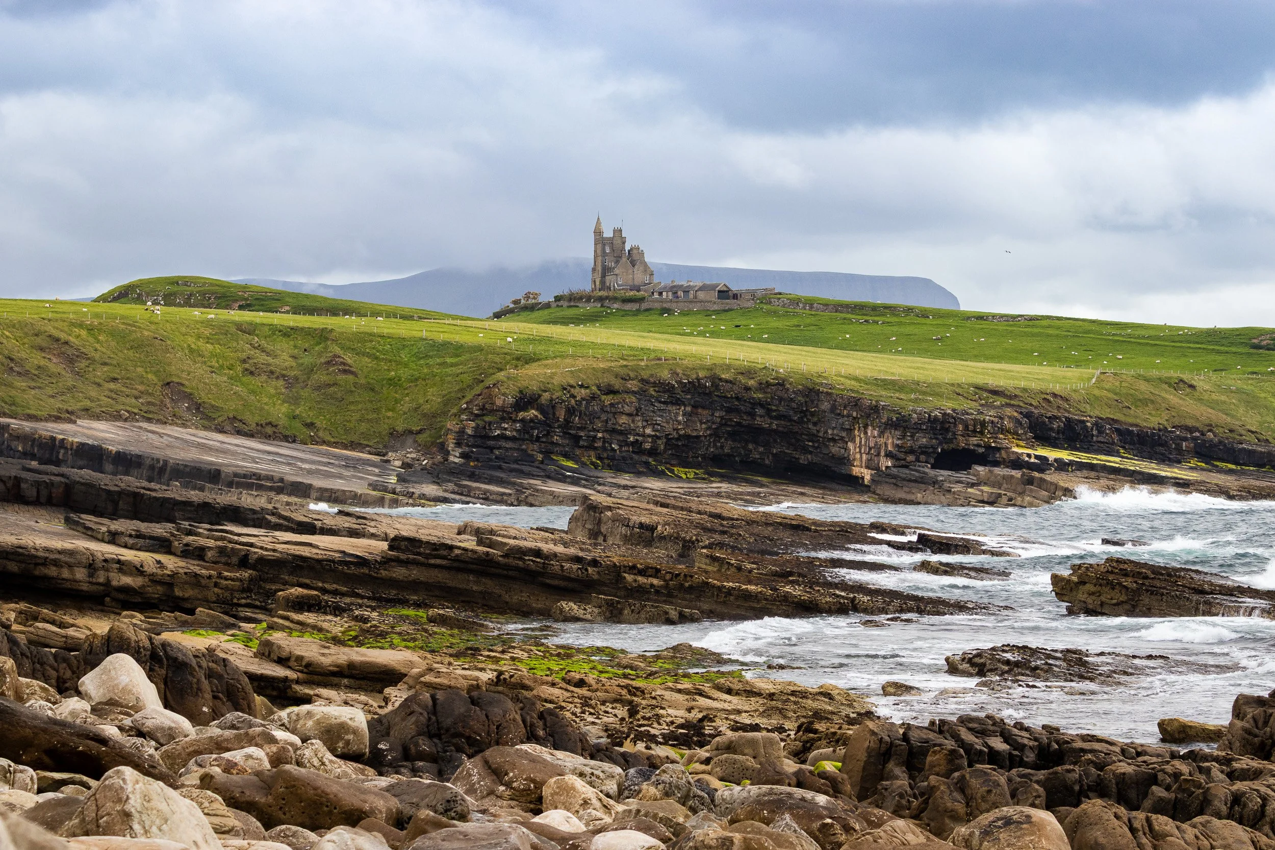 A rocky shoreline with waves crashing against rocks, grassy cliffs in the background, and a castle on a hilltop under a cloudy sky County Sligo.