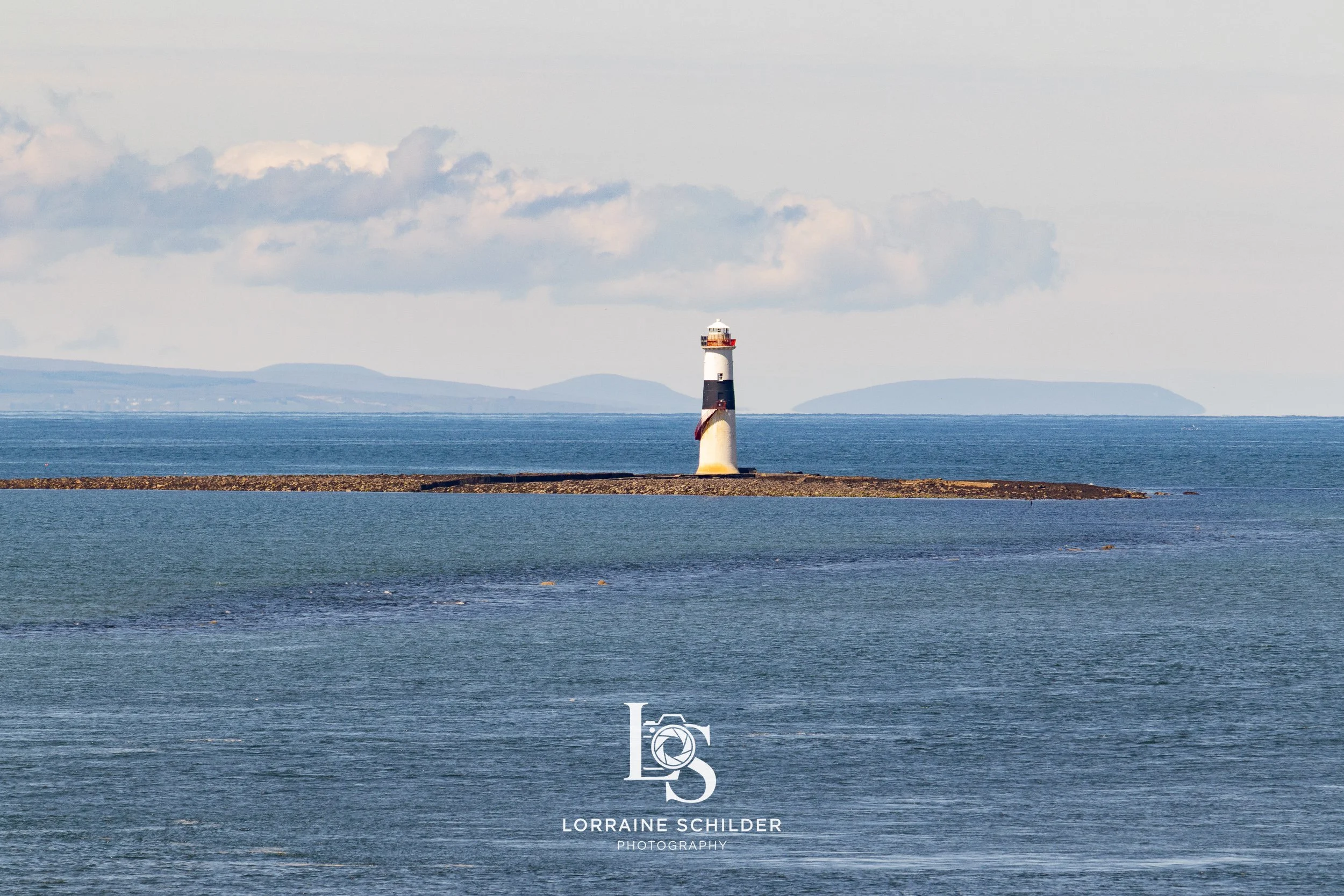 A lighthouse located on a small rocky island in the sea, with distant land and mountains in the background, under a partly cloudy sky. Sligo.