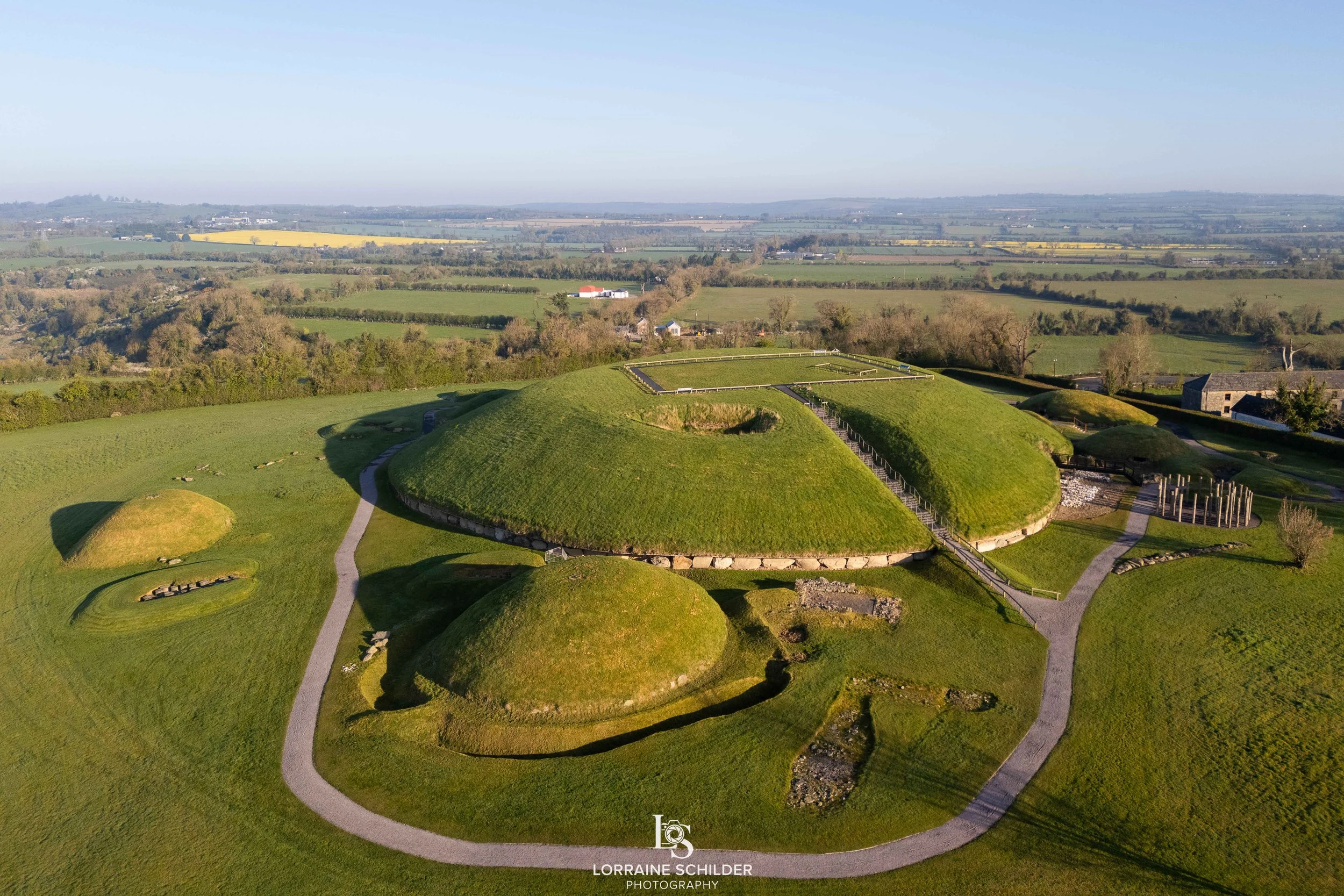 Aerial view of a prehistoric burial mound with grassy domes and pathways, set in a rural landscape with fields and trees in the background. Knowth, Meath.
