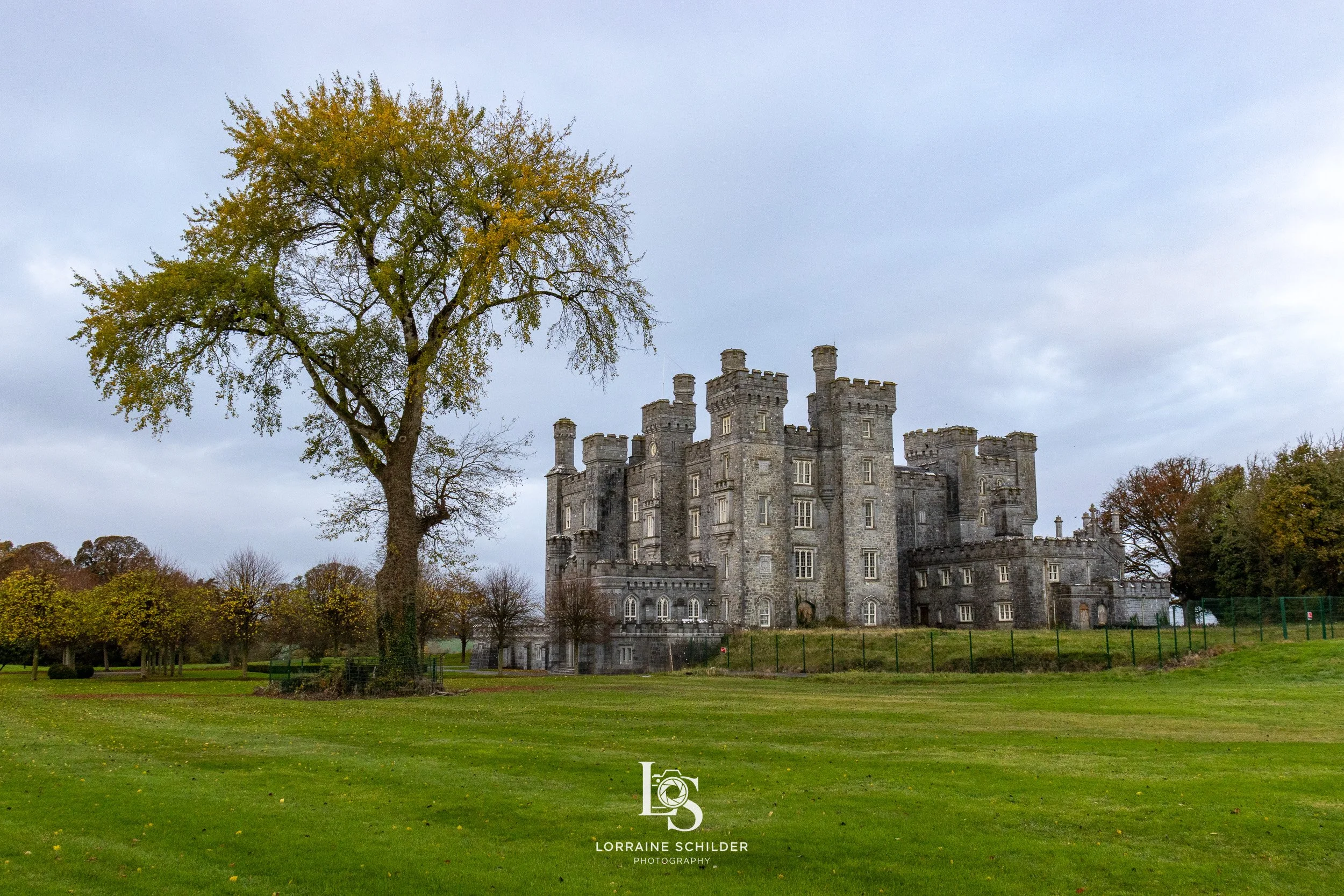 A historic stone castle with turrets and crenellations, surrounded by a grassy lawn and trees with early autumn leaves. Overcast sky in the background, with the photographer's watermark at the bottom. Killeen Castle, Meath.