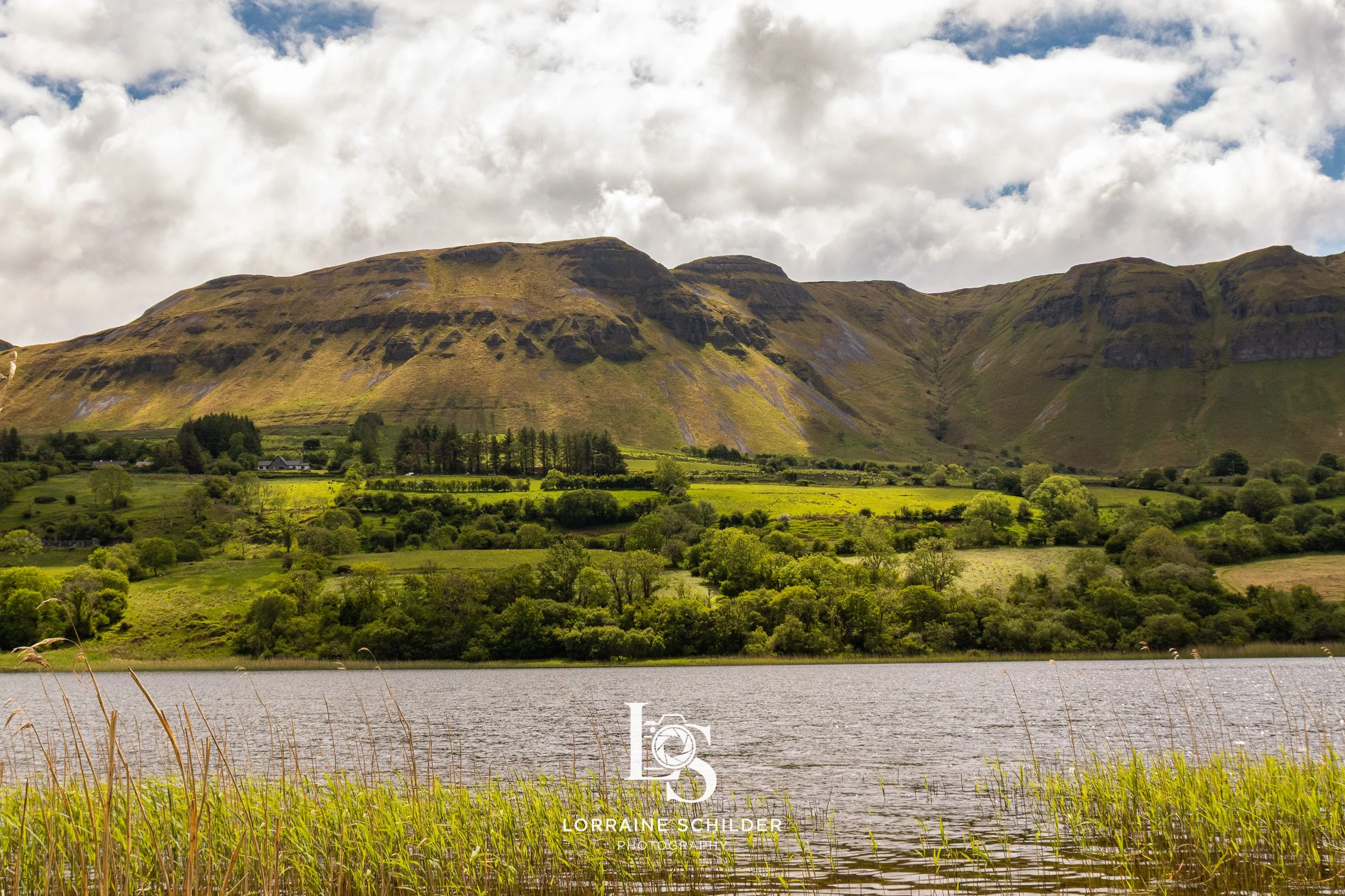 Scenic view of a lake with green grass in the foreground, lush green trees and fields beyond, and large mountains under a partly cloudy sky in the background. Leitrim.