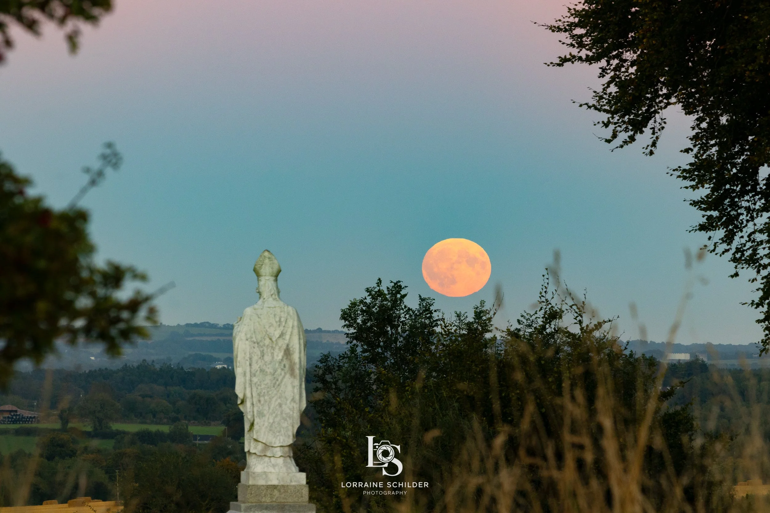 Full moon rising over a scenic landscape with trees, a statue of a figure, and distant hills under a pastel sky.