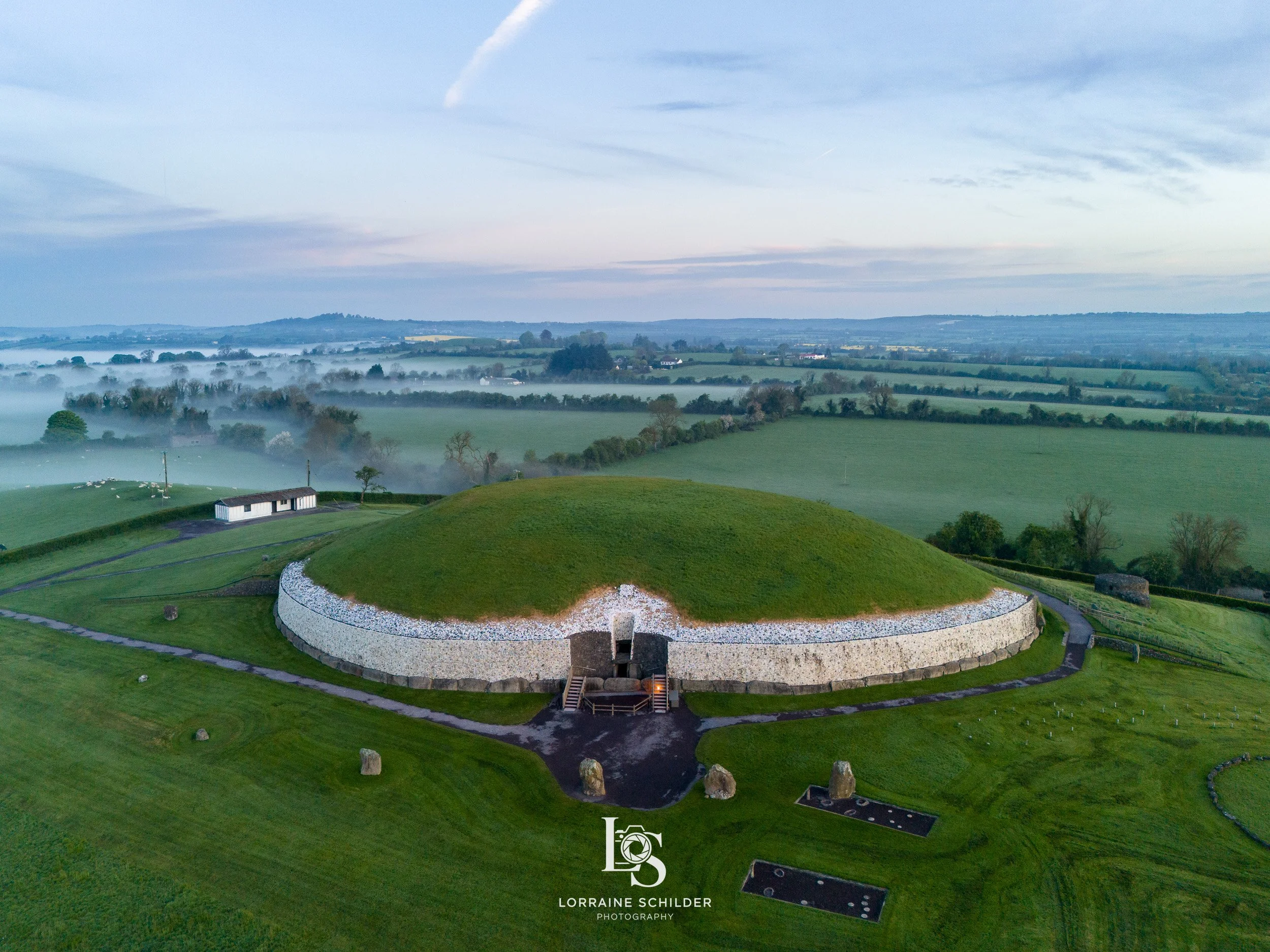 Aerial view of a round, grassy mound surrounded by a white stone wall, with a small entrance and stairs at the front, in a landscape of rolling green fields and scattered trees under a cloudy sky. Newgrange, Meath.