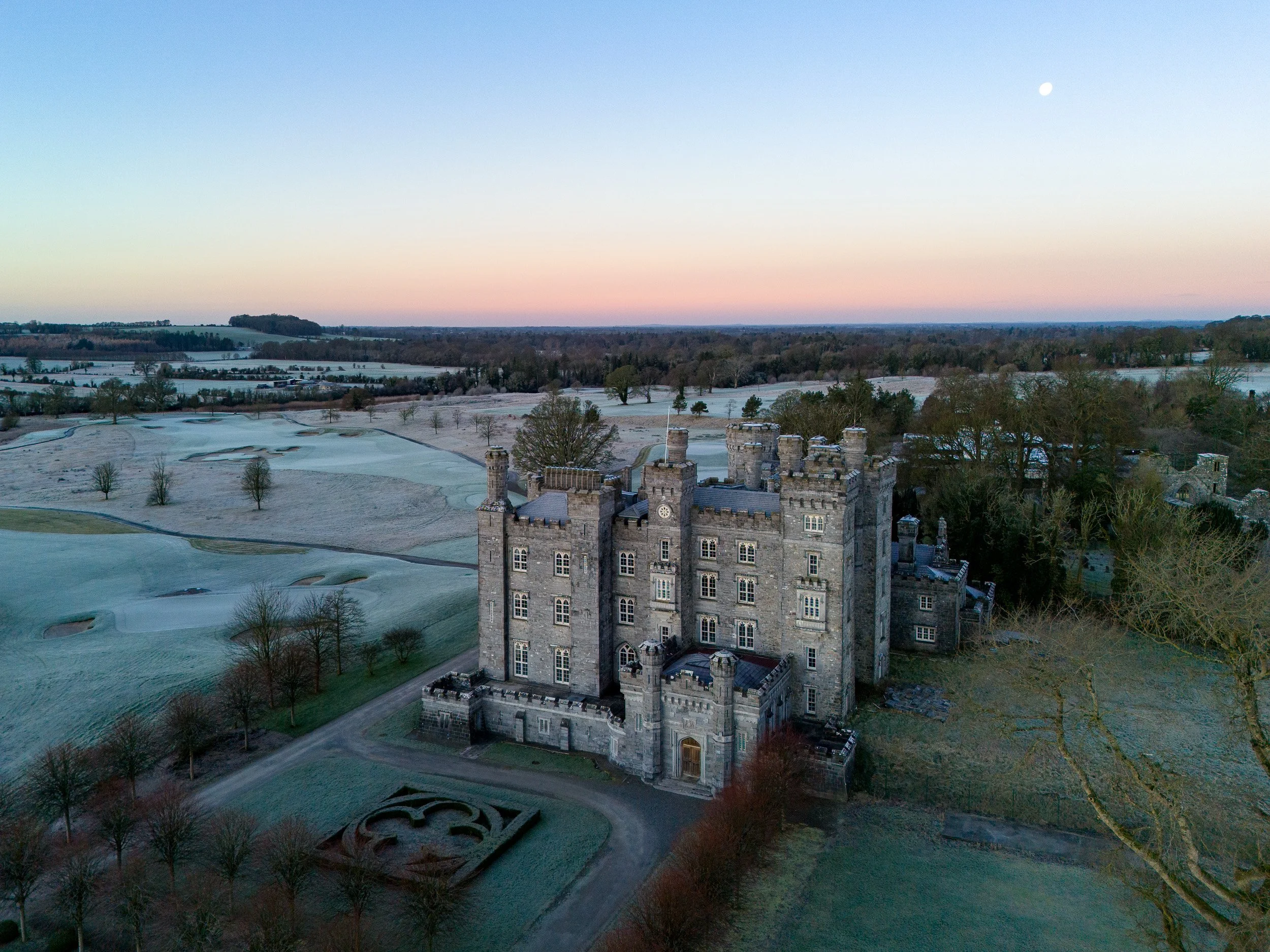 Aerial view of a large historic castle surrounded by frosty landscape and a golf course, under a sky with a visible moon at sunrise.