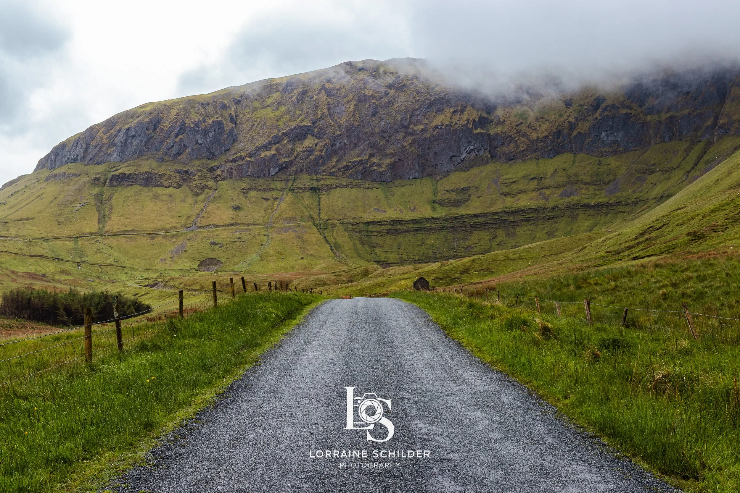 A gravel road leading towards green rolling hills and a mountain in the background, partially covered with mist or low clouds. Sligo.