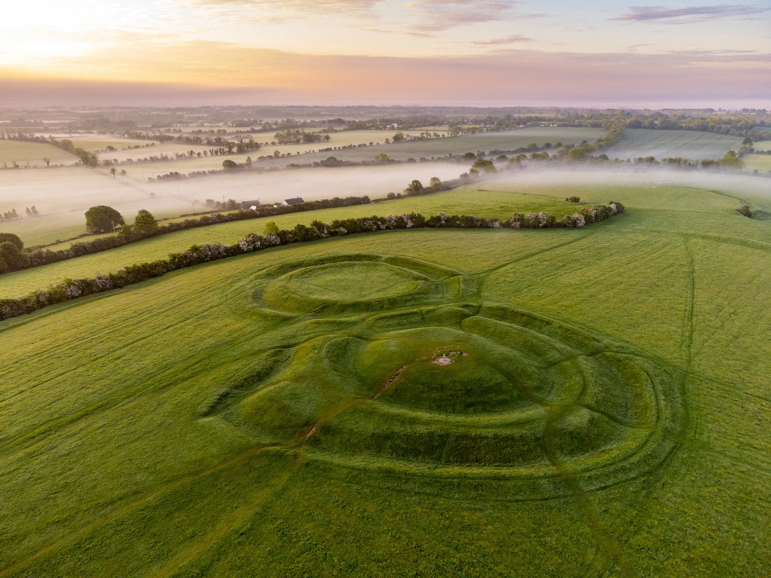 An aerial view of a large green field with a castle shape made of grass, surrounded by trees and small bushes, during sunrise with fog in the distance.