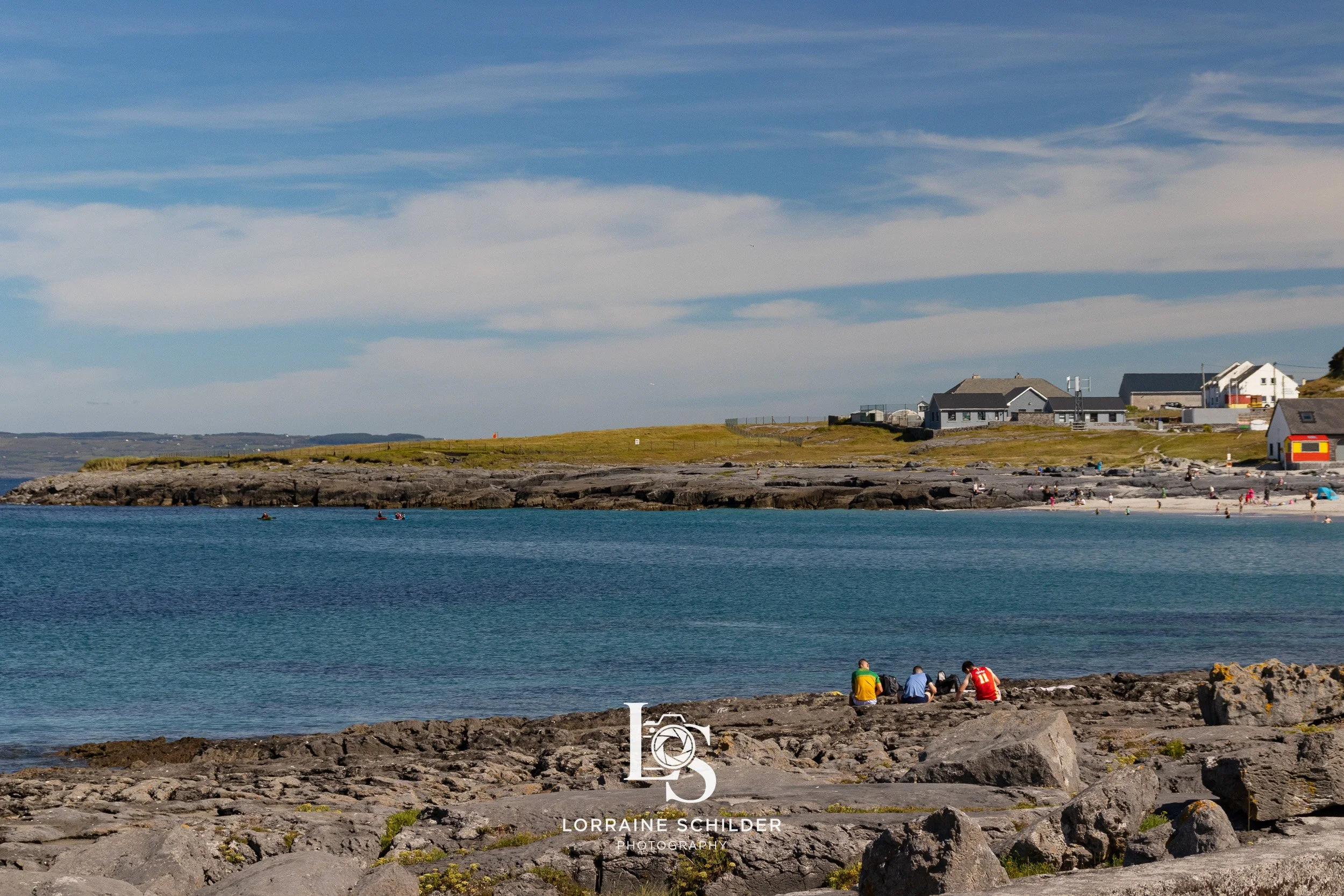 A coastal scene with rocky shoreline, blue water, and a few people sitting on the rocks. In the background, there are buildings and a beach with more people, under a partly cloudy sky.  Inisheer, Galway.