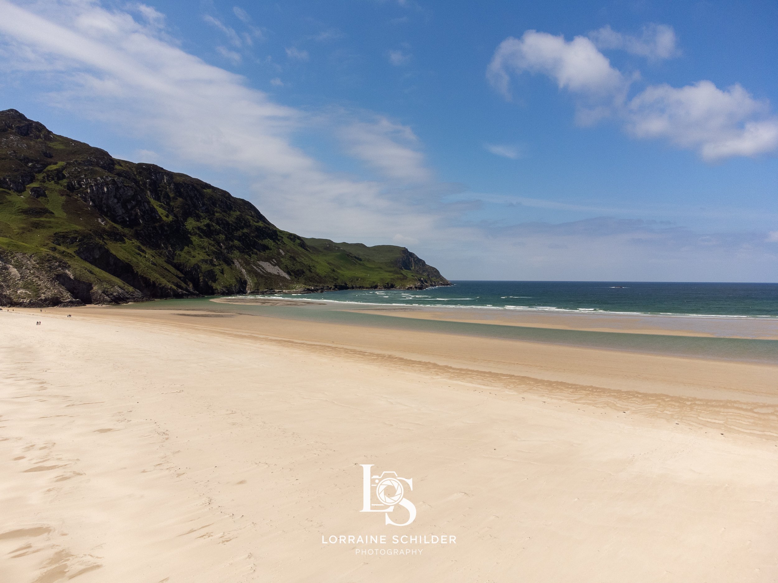 A wide sandy beach with rolling waves, green hillside cliffs on the left, and a partly cloudy blue sky.  Donegal.