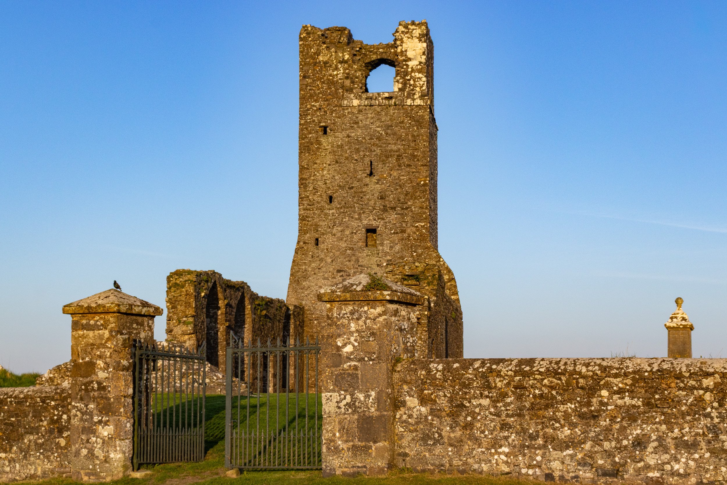Ancient stone tower ruins with a blue sky backdrop. The tower is partially collapsed, with an arched opening near the top. Surrounding stone walls and gate with a bird perched on top are visible in the foreground.