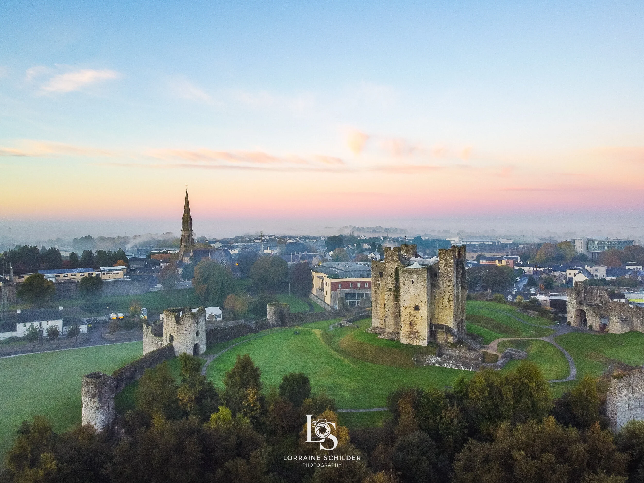 An aerial view Trim Castle on a grassy hill, surrounded by trees, with a small town and church steeple in the background under a pastel sky.