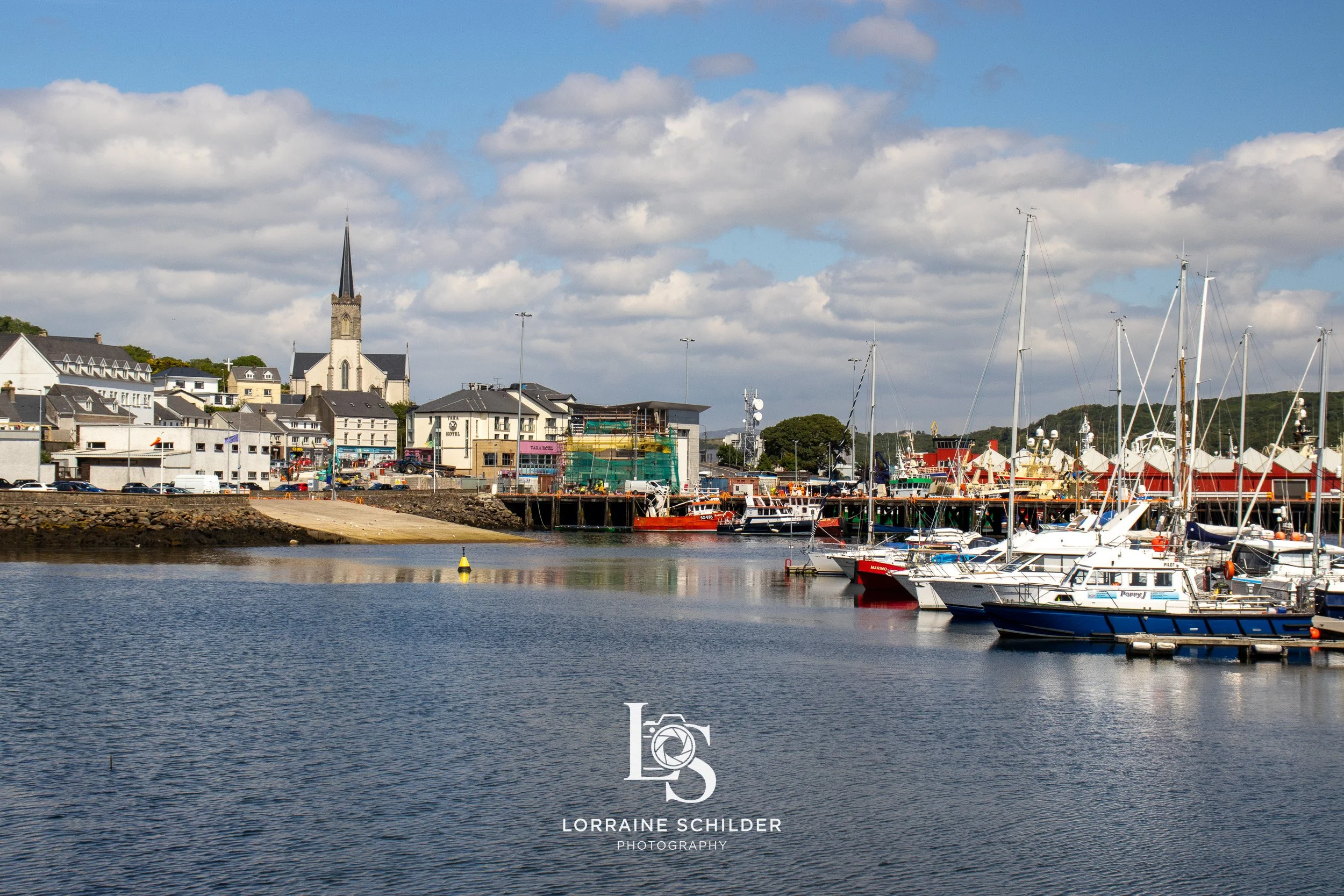 Scenic view of a harbor with boats docked, a town with houses and a church with a tall steeple in the background, and a partly cloudy sky.  Donegal.