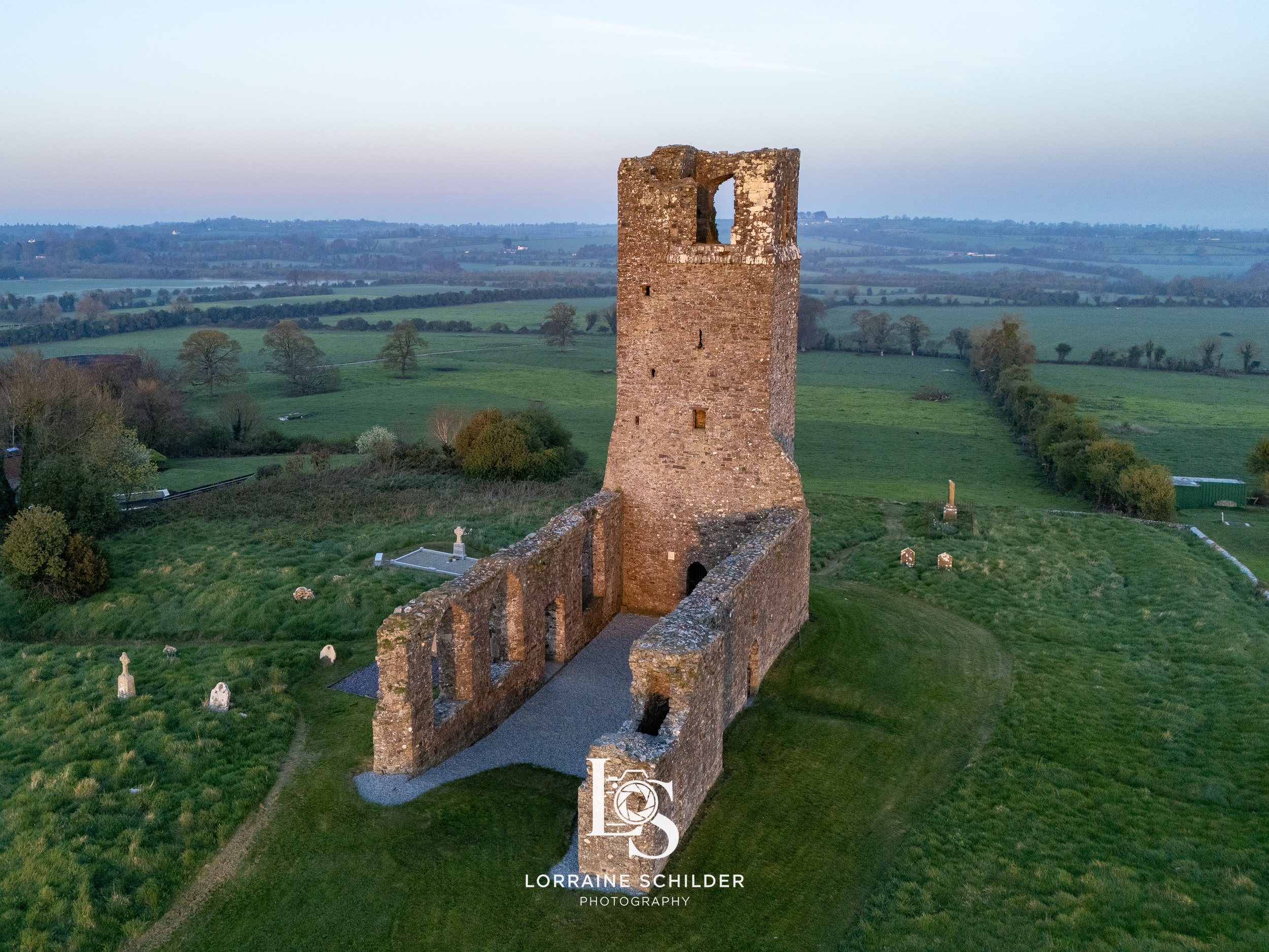 An aerial view of an old stone ruin in a green field with trees and distant hills, taken during sunrise. Skyrne, Meath.