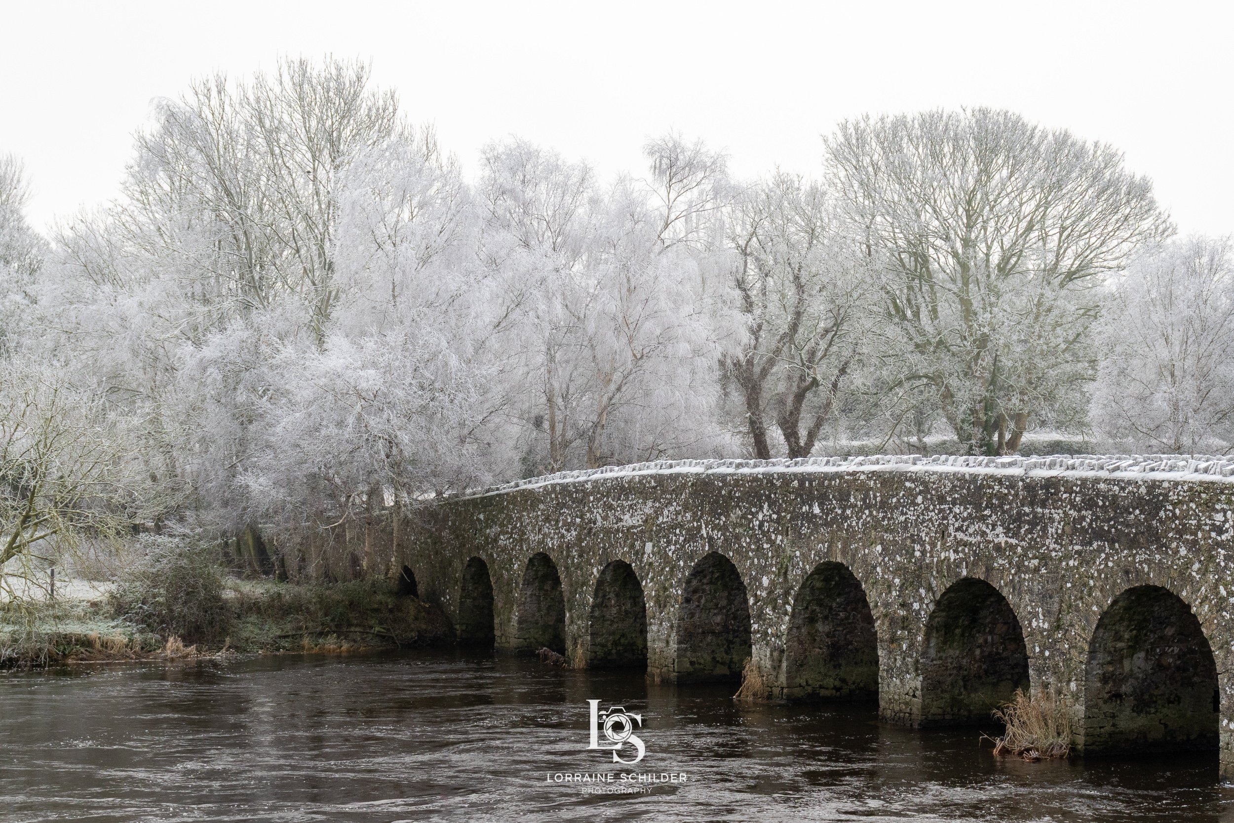 Snow-covered stone bridge over a river, surrounded by leafless trees winter landscape.  Bective Abbey, Meath.