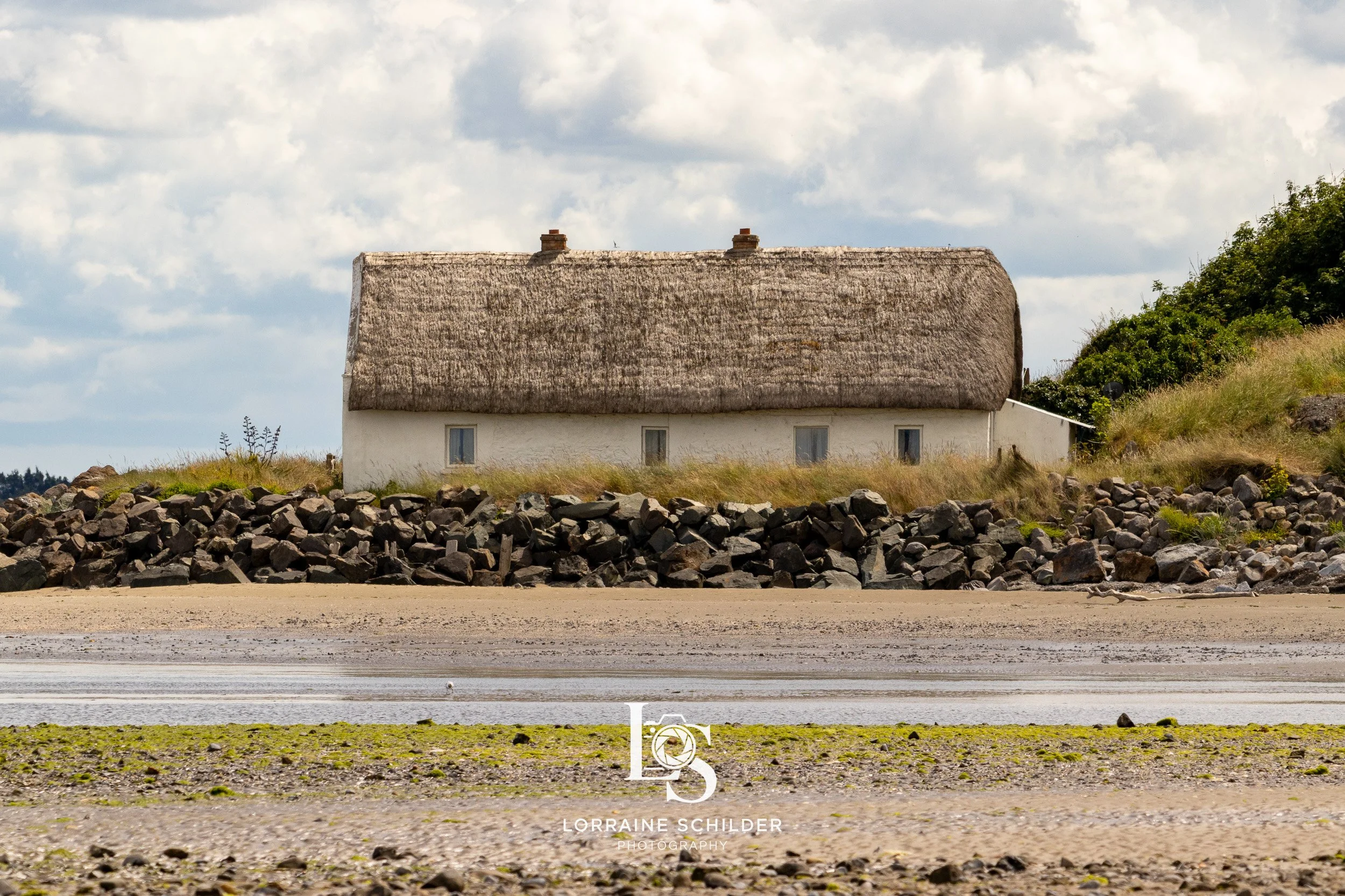 A quaint house with a thatched roof on a rocky shoreline, with grassy terrain and a cloudy sky. Laytown, Meath.