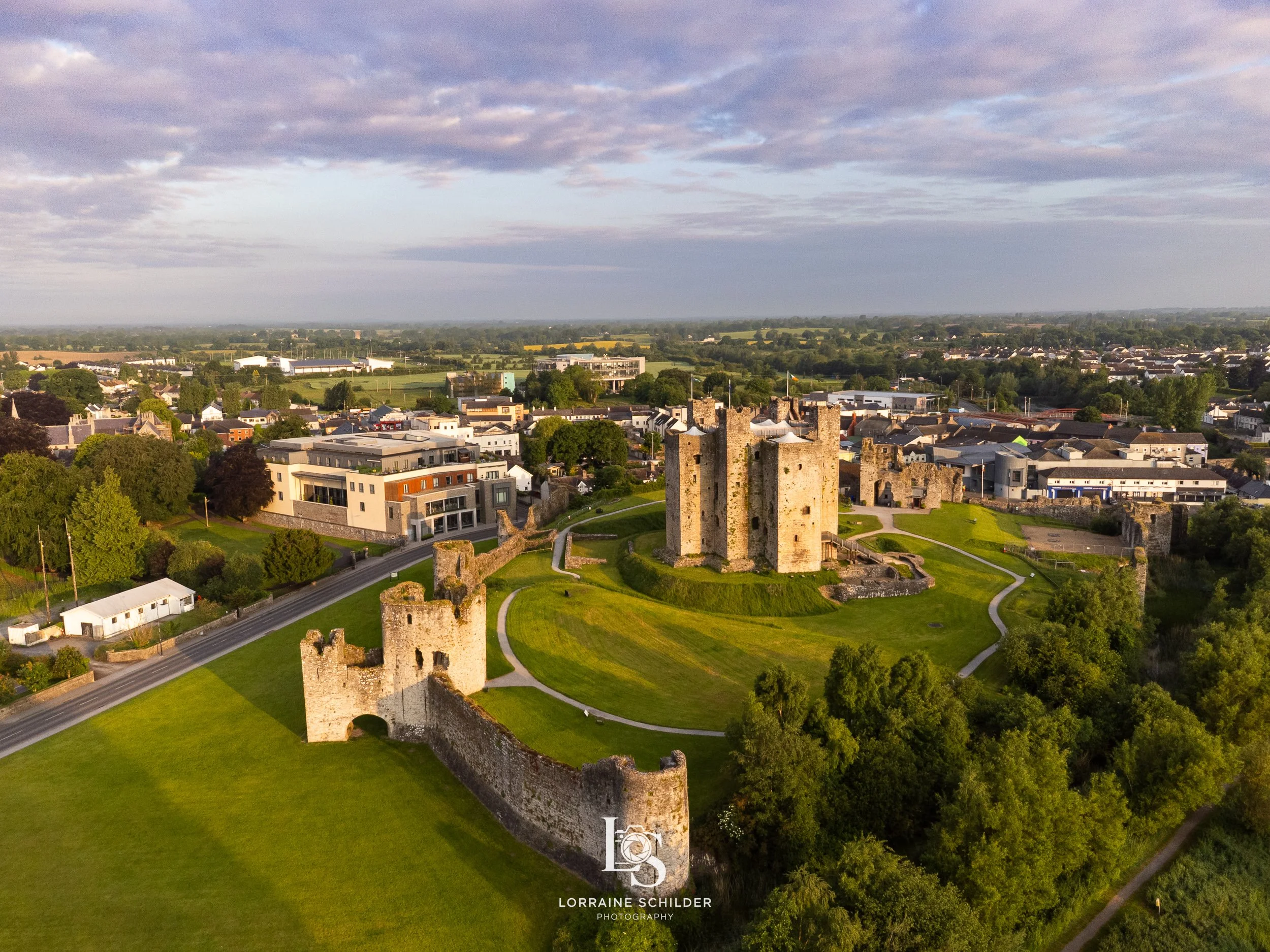 Aerial view of Trim castle ruins surrounded by green lawns and pathways, with a modern town and farmland in the background under a partly cloudy sky.