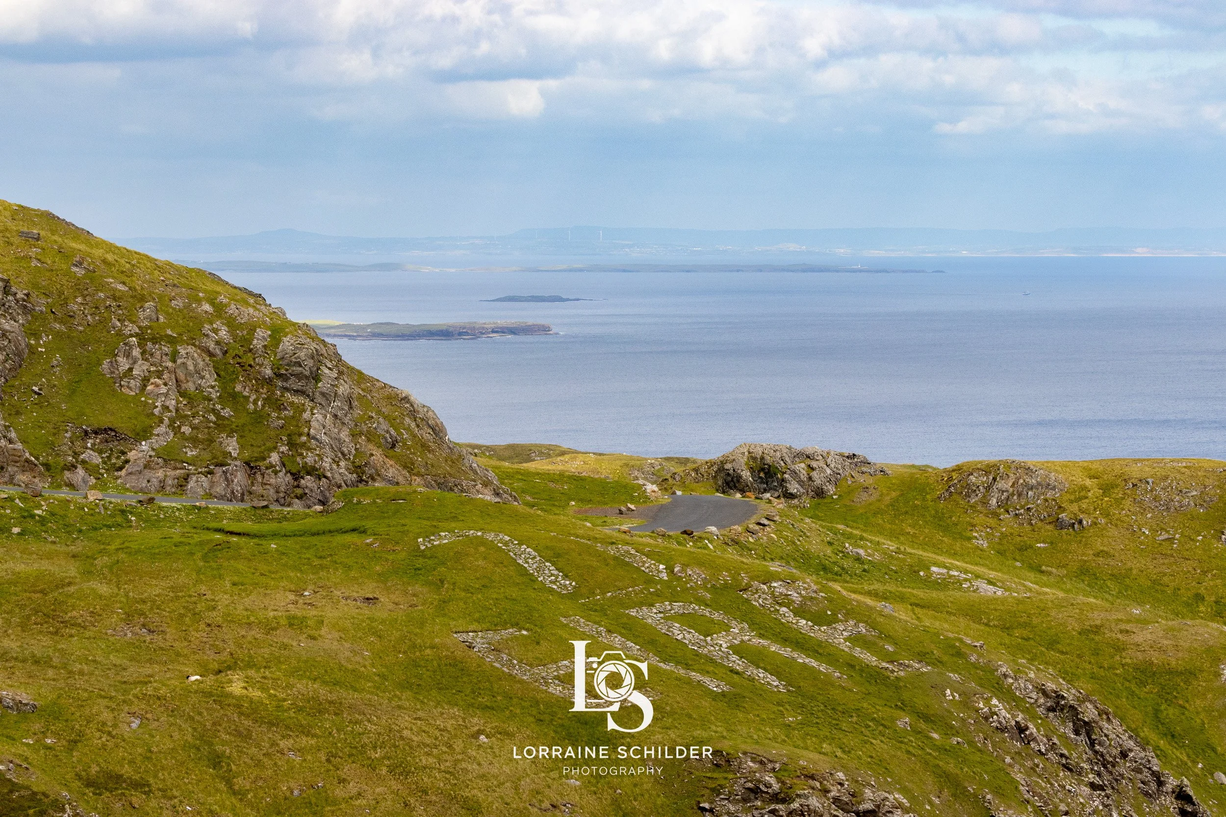 A scenic view of green rolling hills with a hillside inscription carved into the grass, overlooking a large body of water with small islands in the distance.  Donegal.
