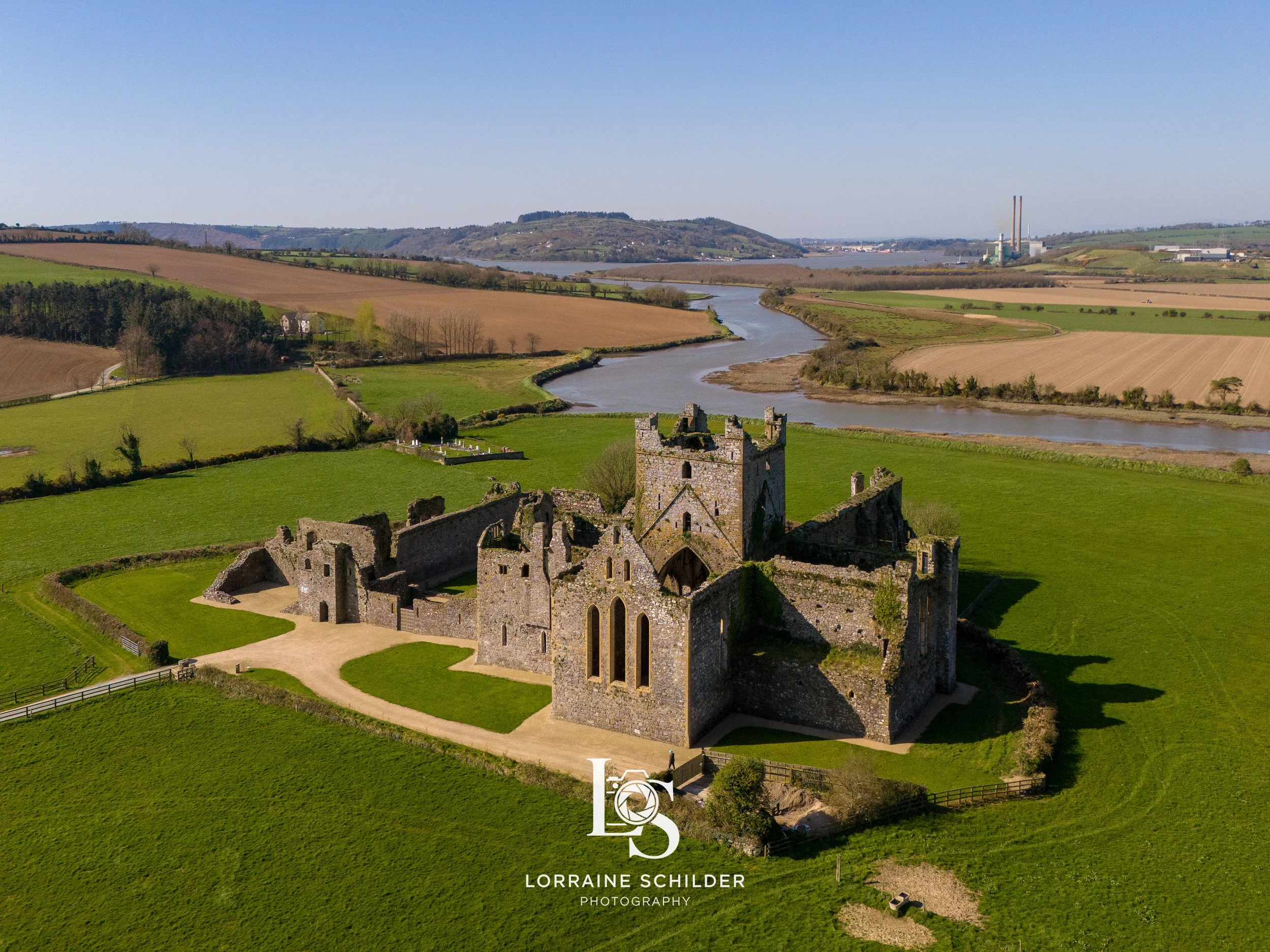 An aerial view of a historic stone castle ruins located on a grassy field, with a river and rolling hills in the background. Wexford.