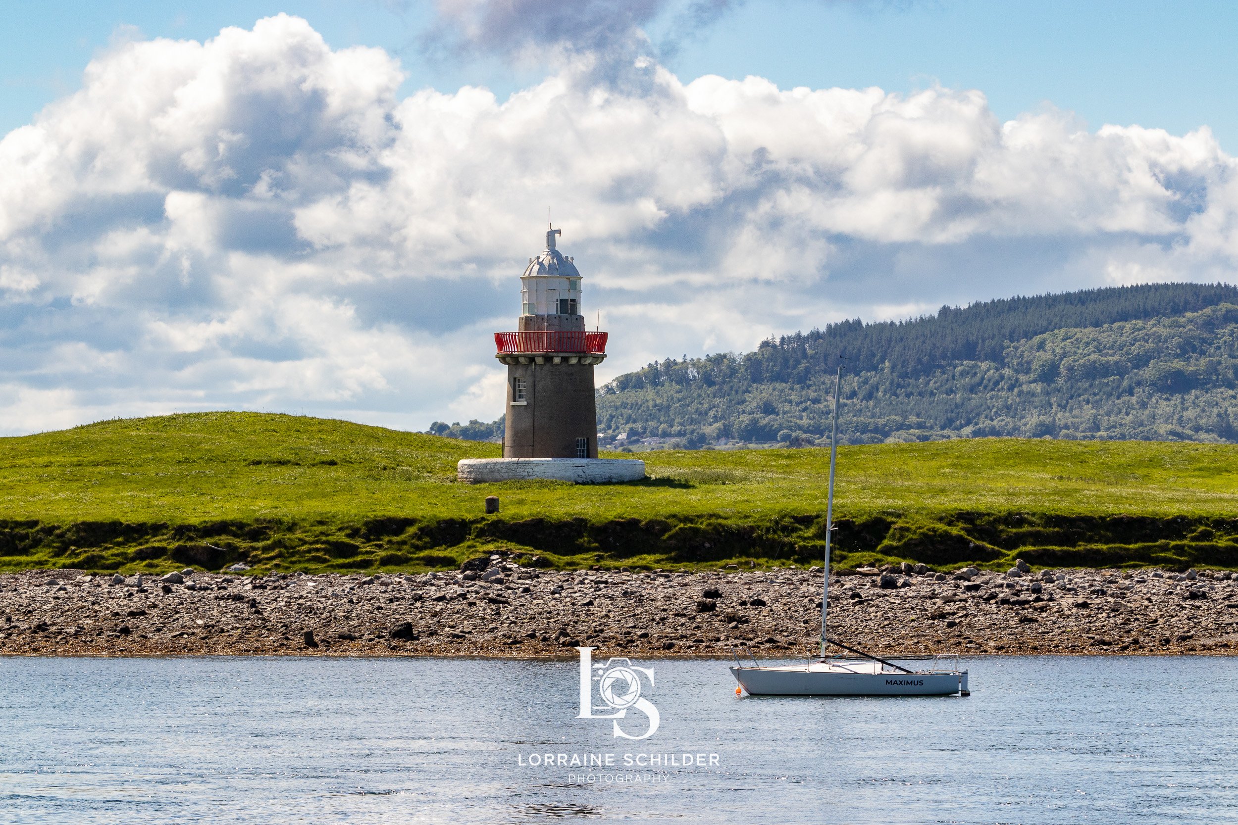 A lighthouse on a grassy hill beside a body of water with a sailboat in the foreground. Sligo.