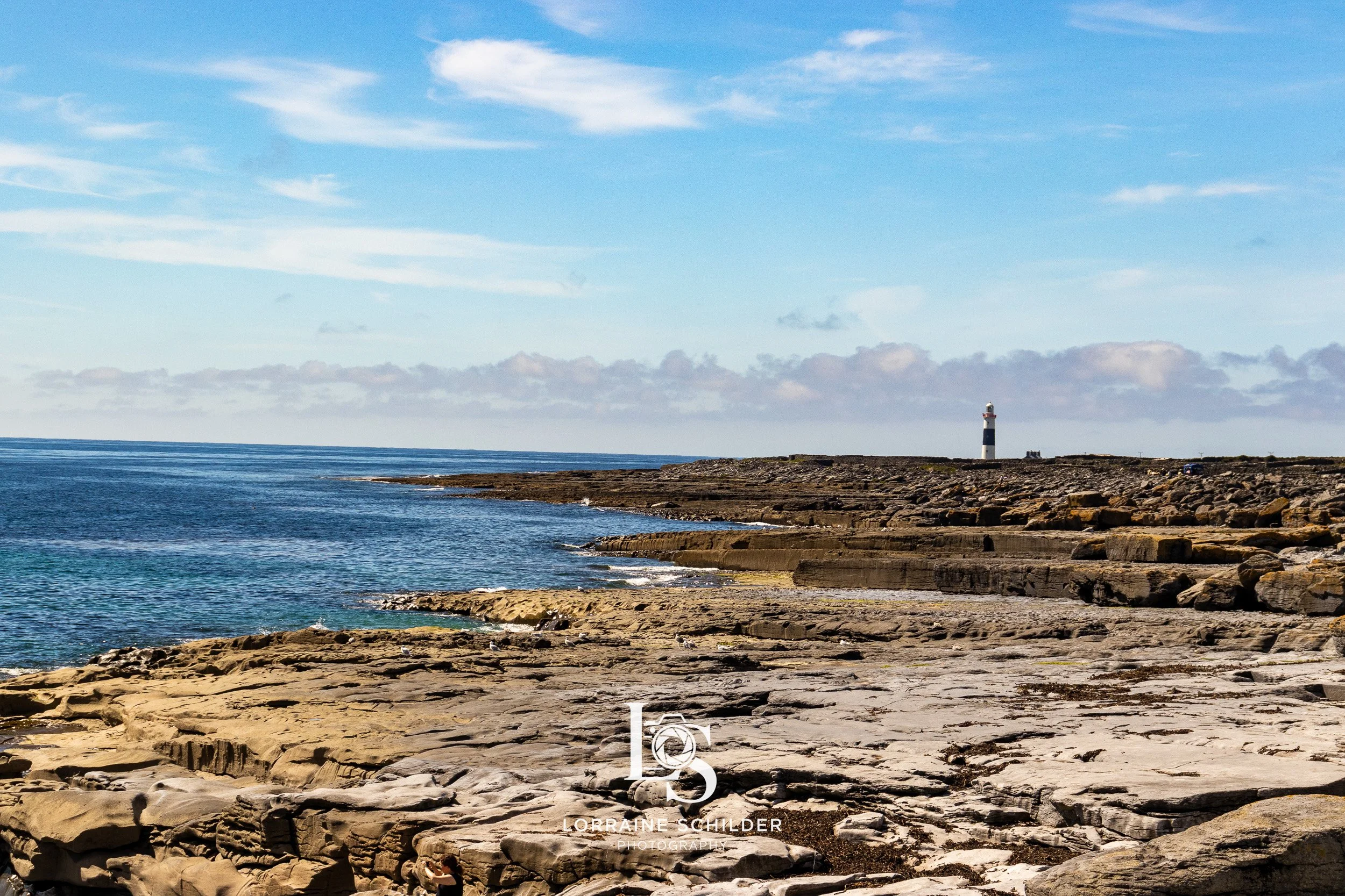 A rocky coastline with a lighthouse in the distance, under a partly cloudy sky.  Inisheer, Galway.