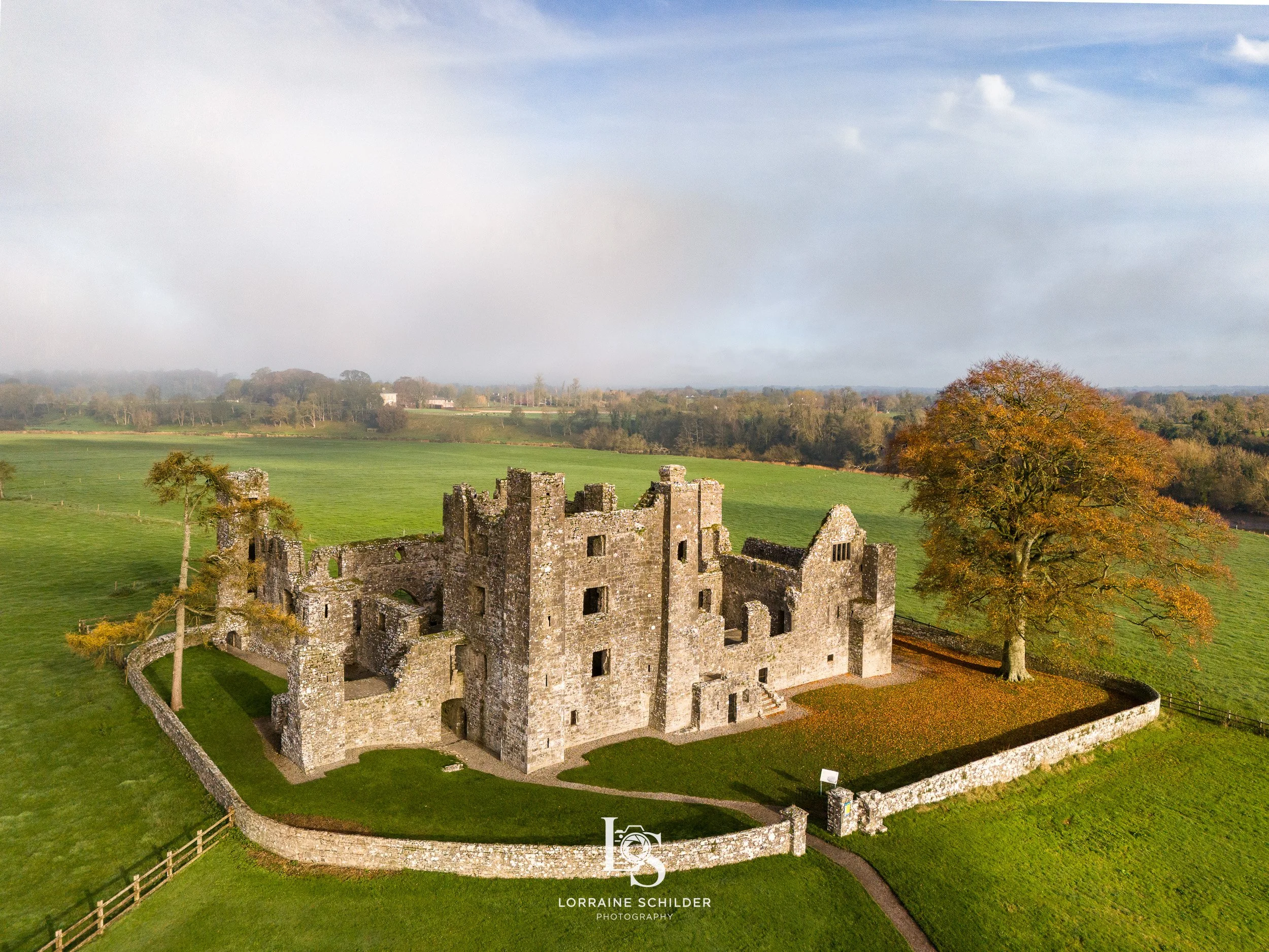 Aerial view of an ancient stone castle surrounded by a green lawn and trees with autumn foliage, with open fields and a cloudy sky in the background.