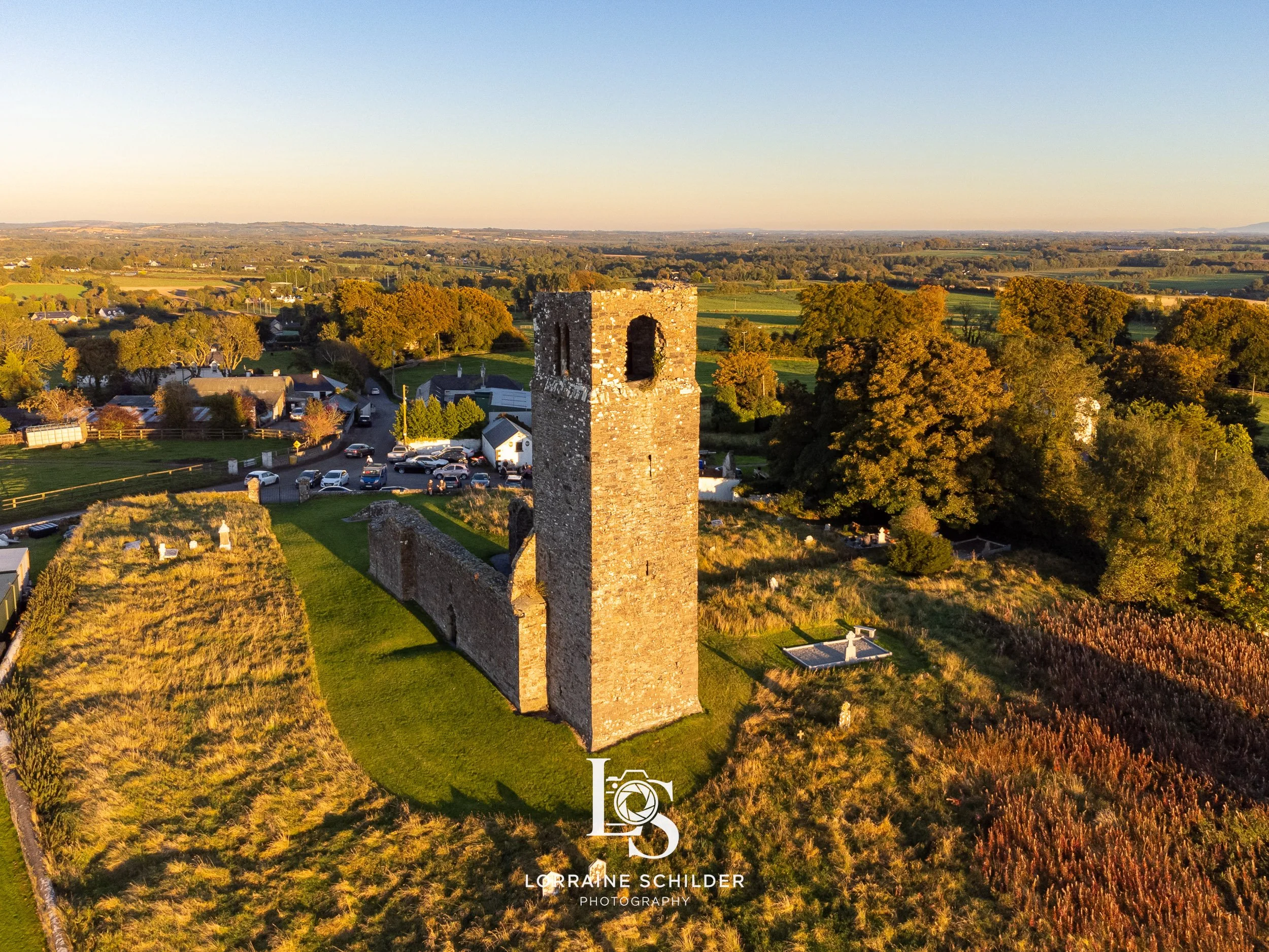 Aerial view of an ancient stone tower surrounded by green fields, trees, and a few houses at sunset. Skyrne, Meath.