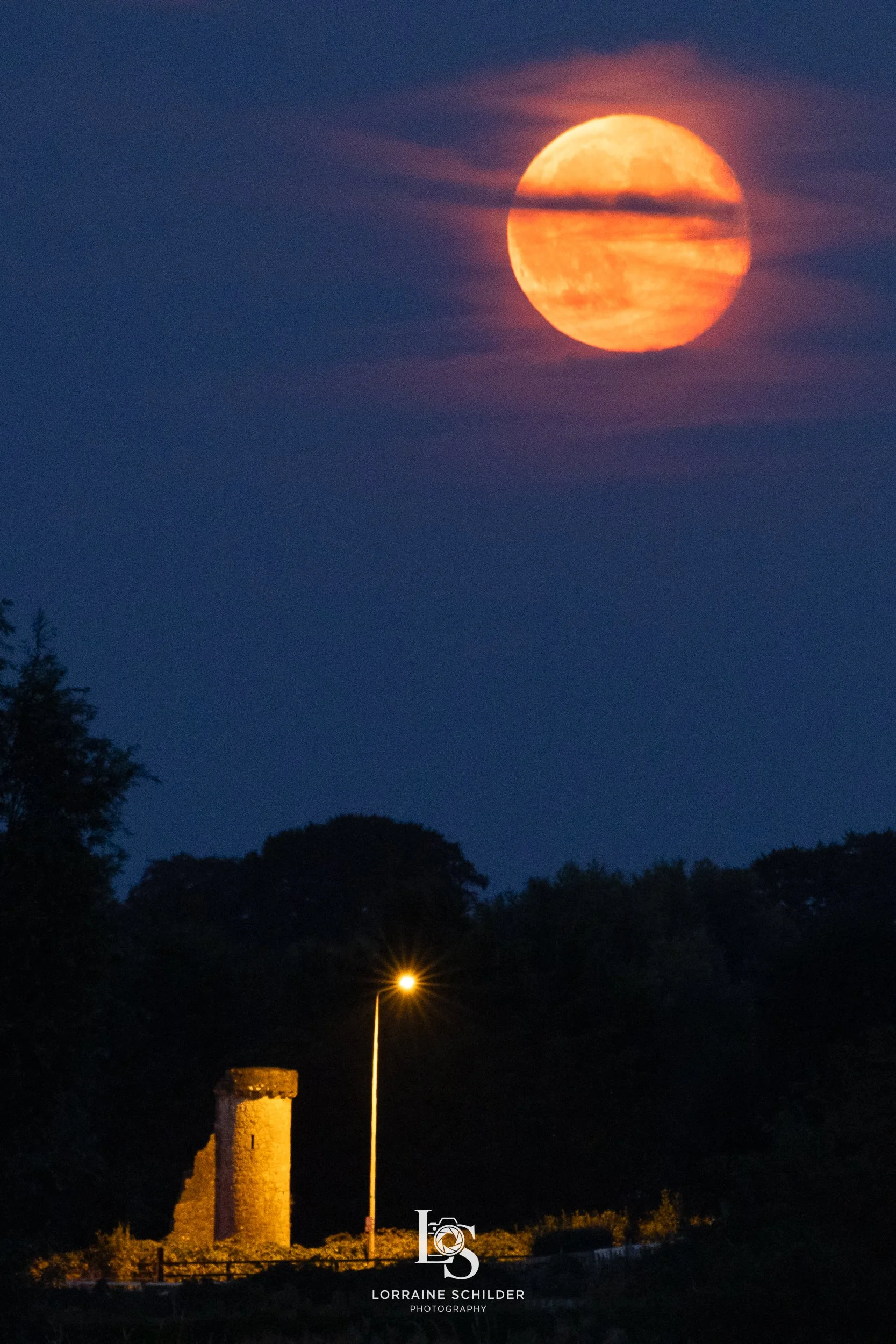 A large orange moon rising in a dark blue sky, with some clouds partially obscuring it. At the bottom, there is a stone tower, a streetlamp, and some trees silhouetted against the dark landscape.