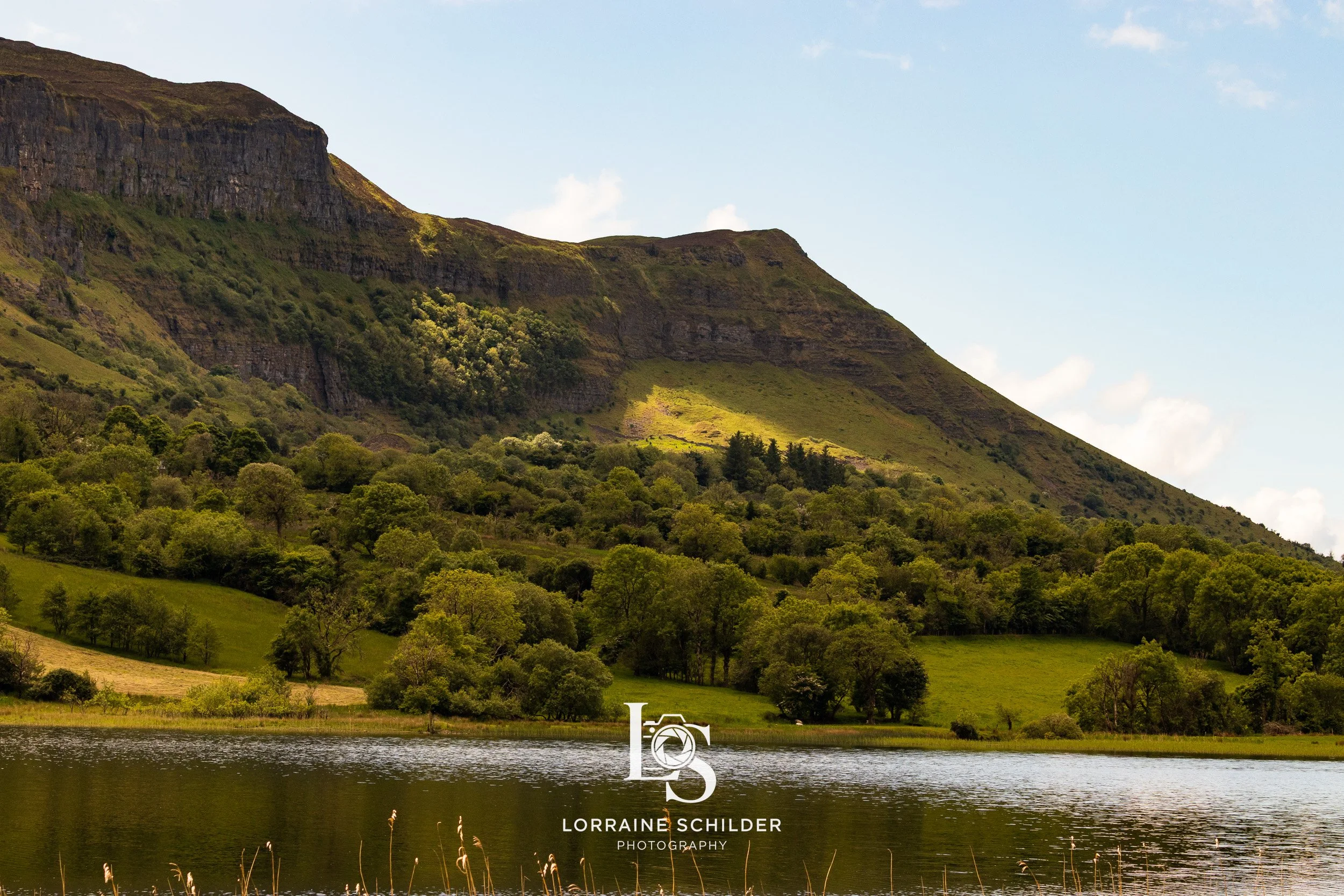 A scenic landscape of green hills and a mountain with a lake in the foreground. The sky is mostly clear with a few clouds. Leitrim.