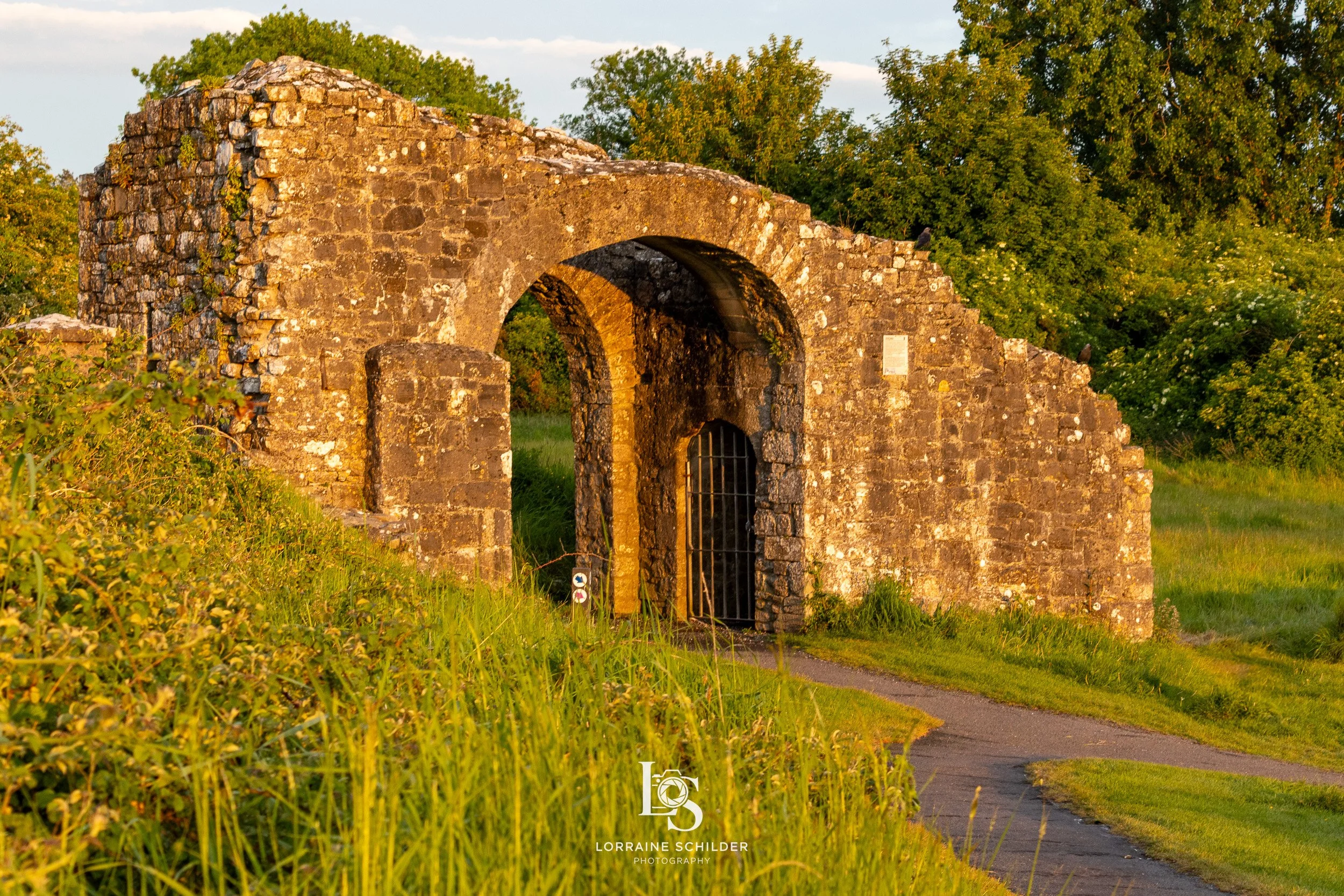 Ancient stone archway with a small barred gate leading through it, set in a grassy landscape with trees in the background, illuminated by warm golden sunlight.
