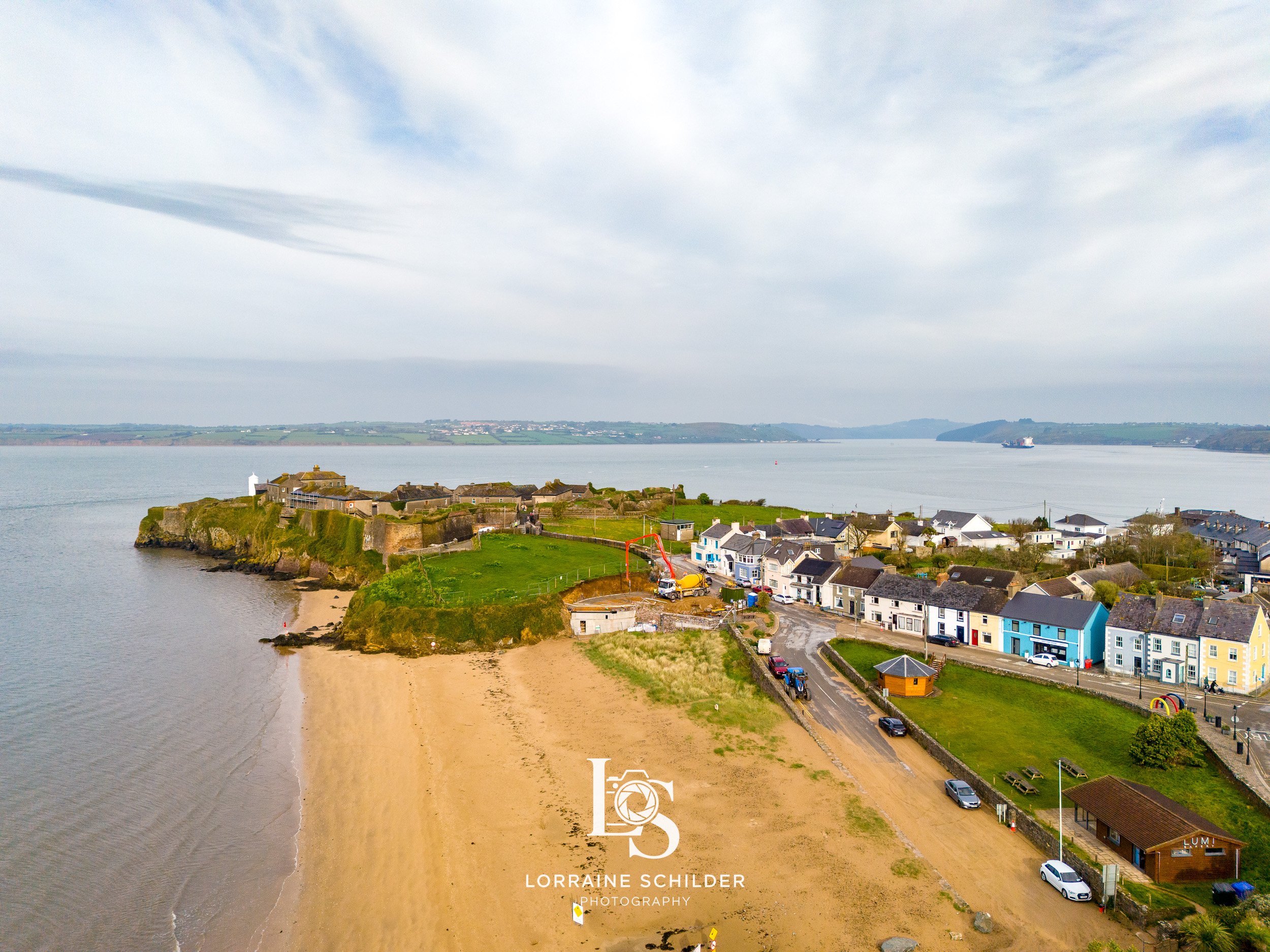 Aerial view of a coastal town with a sandy beach, colorful houses, and a historical fort on a hill, with water and boats in the background. Wexford.