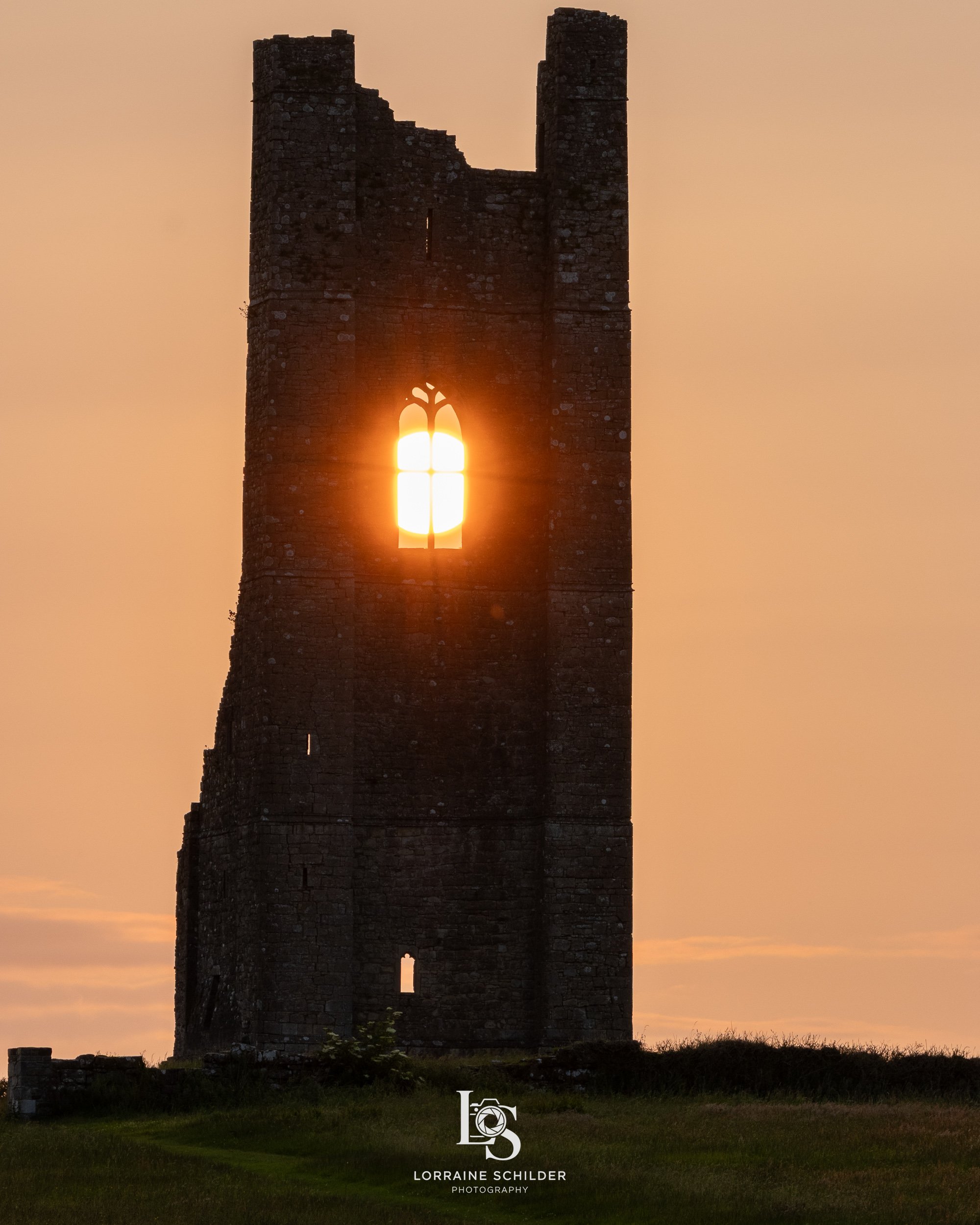 Sunset shines through a tall, ancient stone castle tower with an arched window, creating a warm glow and silhouette effect.