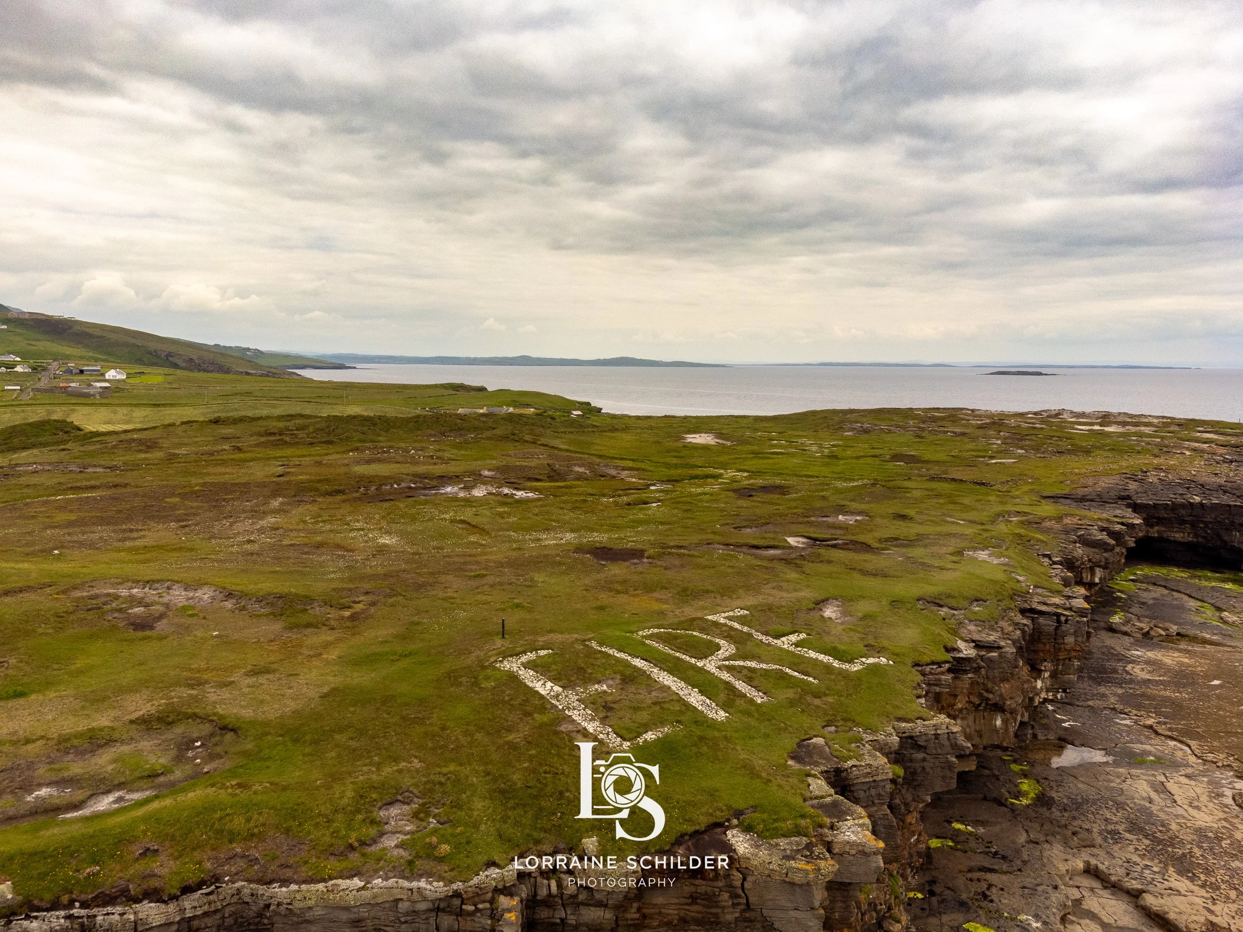 Large green grassy area on a rocky cliffside overlooking the ocean with the word 'Eire' spelled out in white rocks, cloudy sky in the background.  Donegal.
